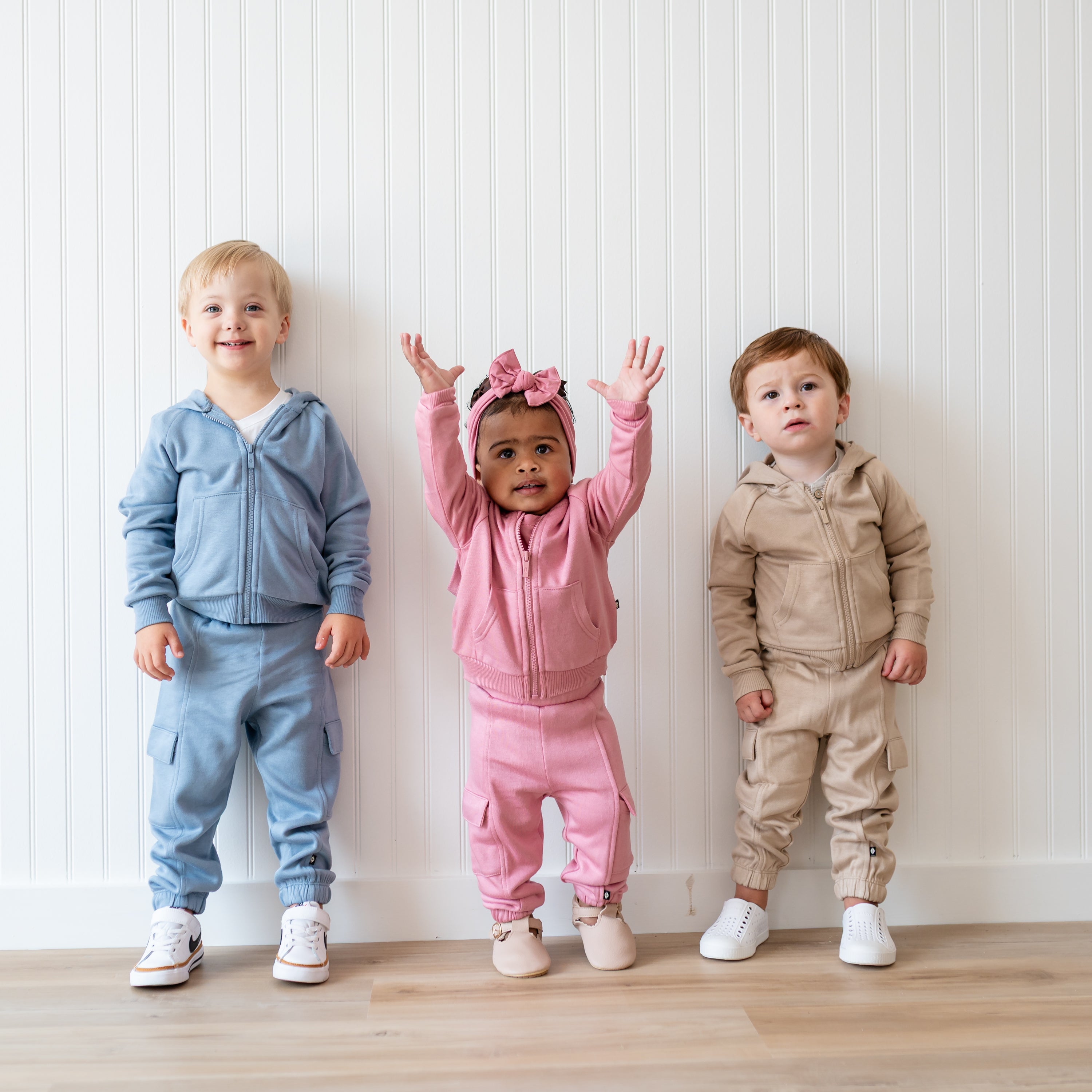 Group of three young children standing in front of a white paneled wall standing beside one another wearing the Fleece Cargo Pants in Slate, Apple Blossom and Almond with the matching fleece zip hoodies