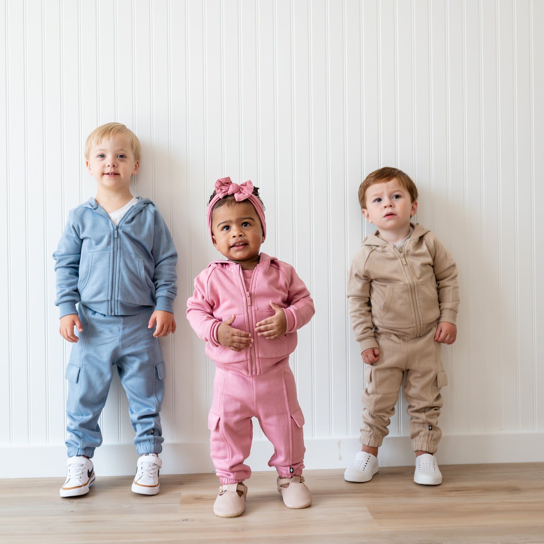 Group of three young children models standing side by side in front of a white paneled wall wearing the Fleece Zip Up Hoodie in Almond, Apple Blossom and Slate with matching Fleece Cargo pants