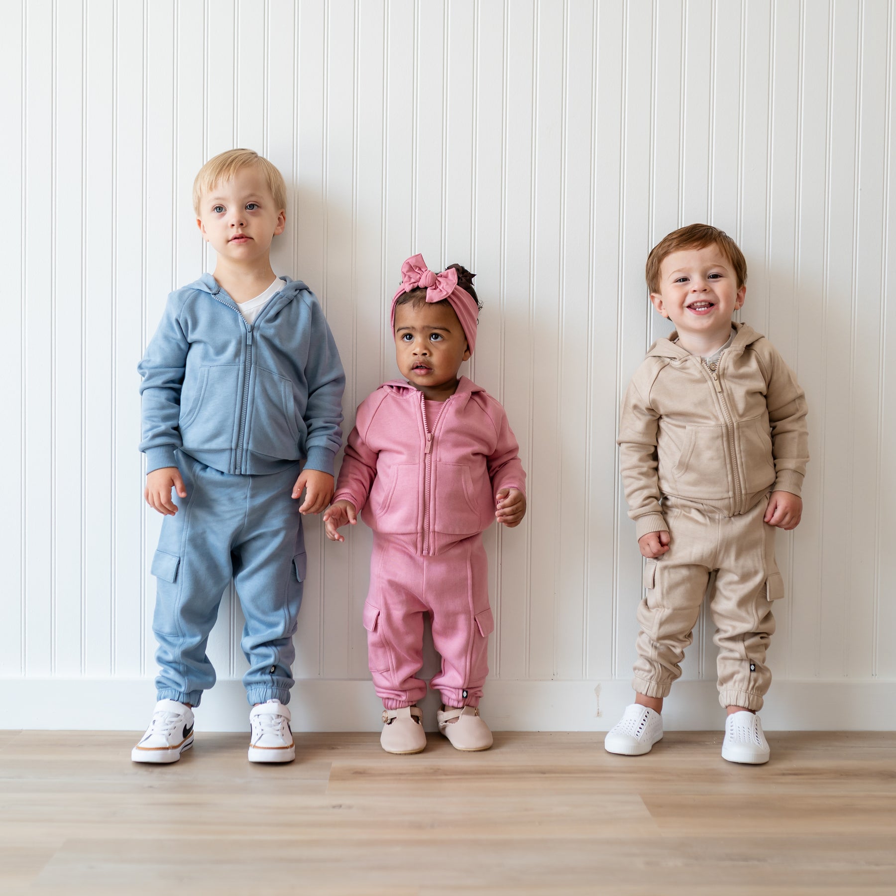 Three young children models standing side by side in front of a white paneled wall wearing the Fleece Zip Up Hoodie in Slate, Apple Blossom and Almond with matching fleece cargo pants