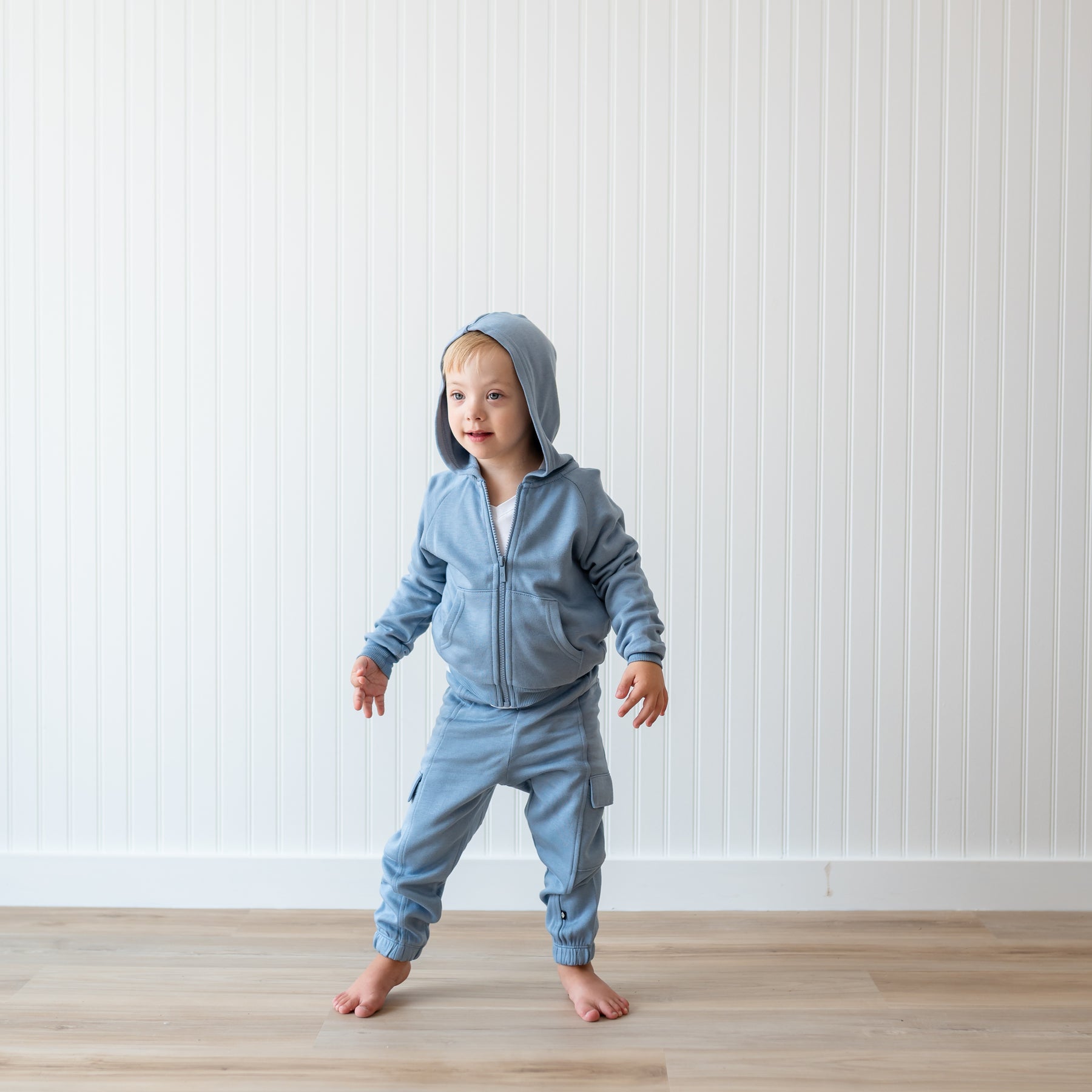 Young boy standing in front of a white paneled wall wearing the Fleece Zip Up Hoodie in Slate and matching fleece cargo pant