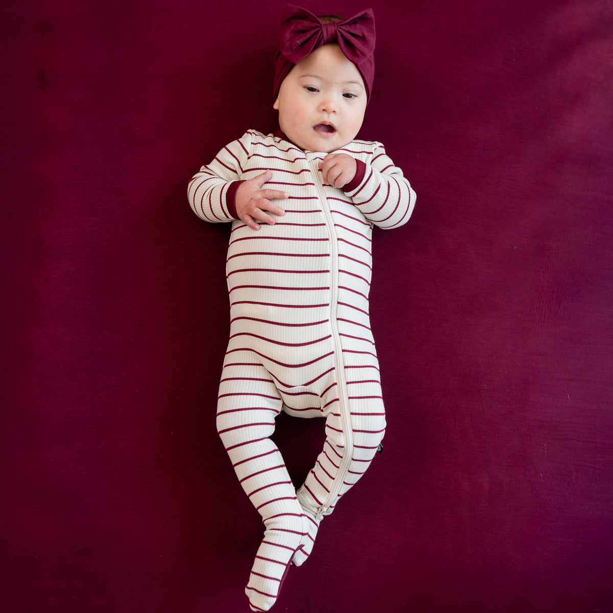 Infant laying on a Burgundy crib sheet wearing the Ribbed Zipper Footie in Burgundy Stripe with burgundy bow headband