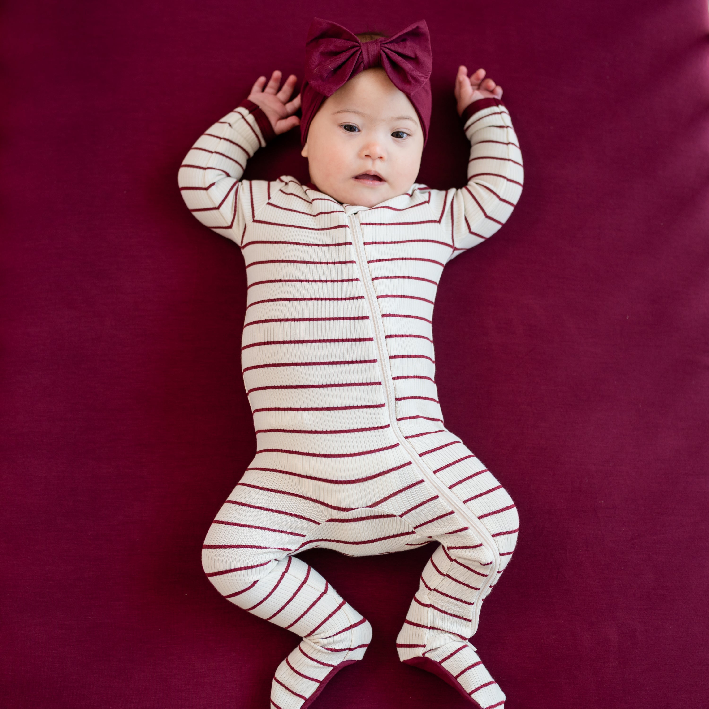 infant laying on a Burgundy crib sheet with arms up and legs bent wearing the Ribbed Zipper Footie in Burgundy Stripe and Burgundy bow headband