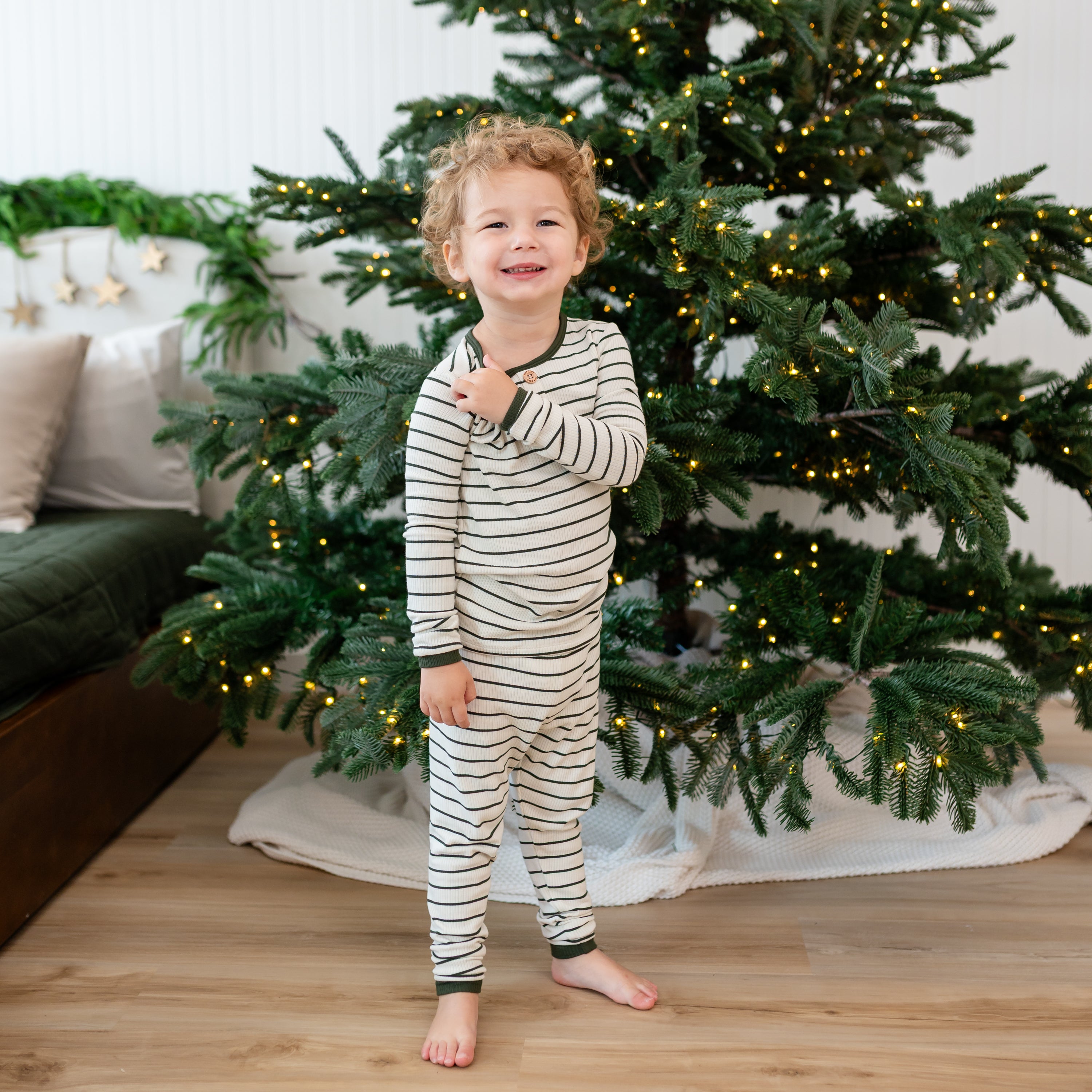 Young toddler standing in front of a lit Christmas Tree wearing the Ribbed Henley Set in Fir Stripe