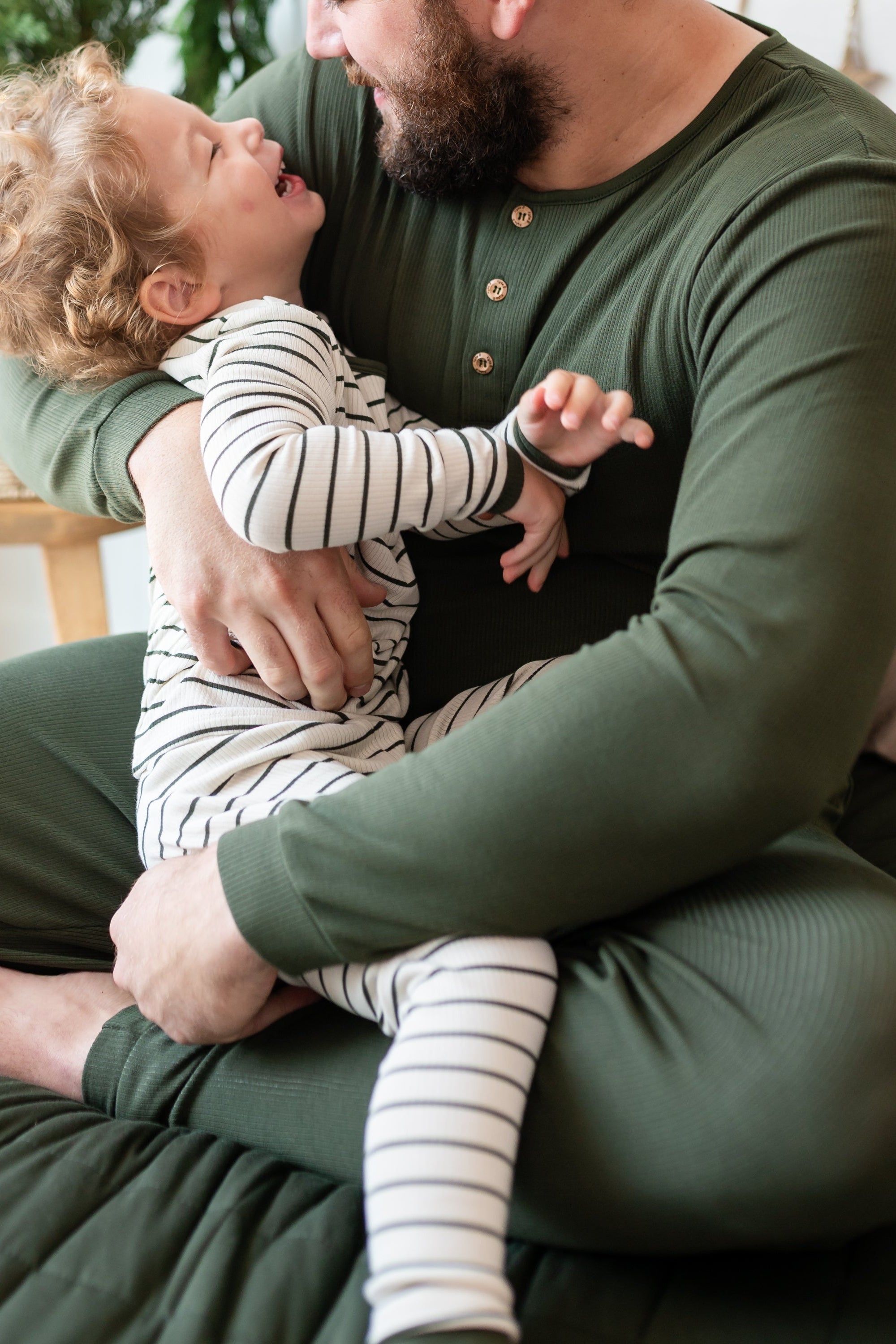 Father wearing the Men's Ribbed Henley Set in Fir sitting on a bed holding his toddler son who is wearing the ribbed henley set in Fir Stripe