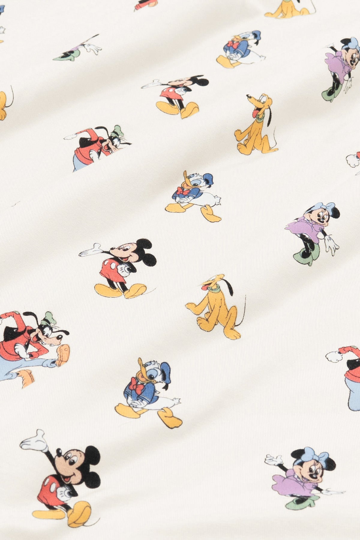 Close up of bamboo fabric swatch: An all-over print featuring: Mickey Mouse in his classic red shorts with white buttons and yellow shoes; Minnie Mouse wearing a blue headbow, a pink dress, and green heels; Goofy jumping in the air and wearing a green hat, a red shirt, blue pants, and orange shoes; Pluto wearing a green collar; and Donald Duck in his classic blue sailor outfit and hat, over a warm grey background color called Oat.