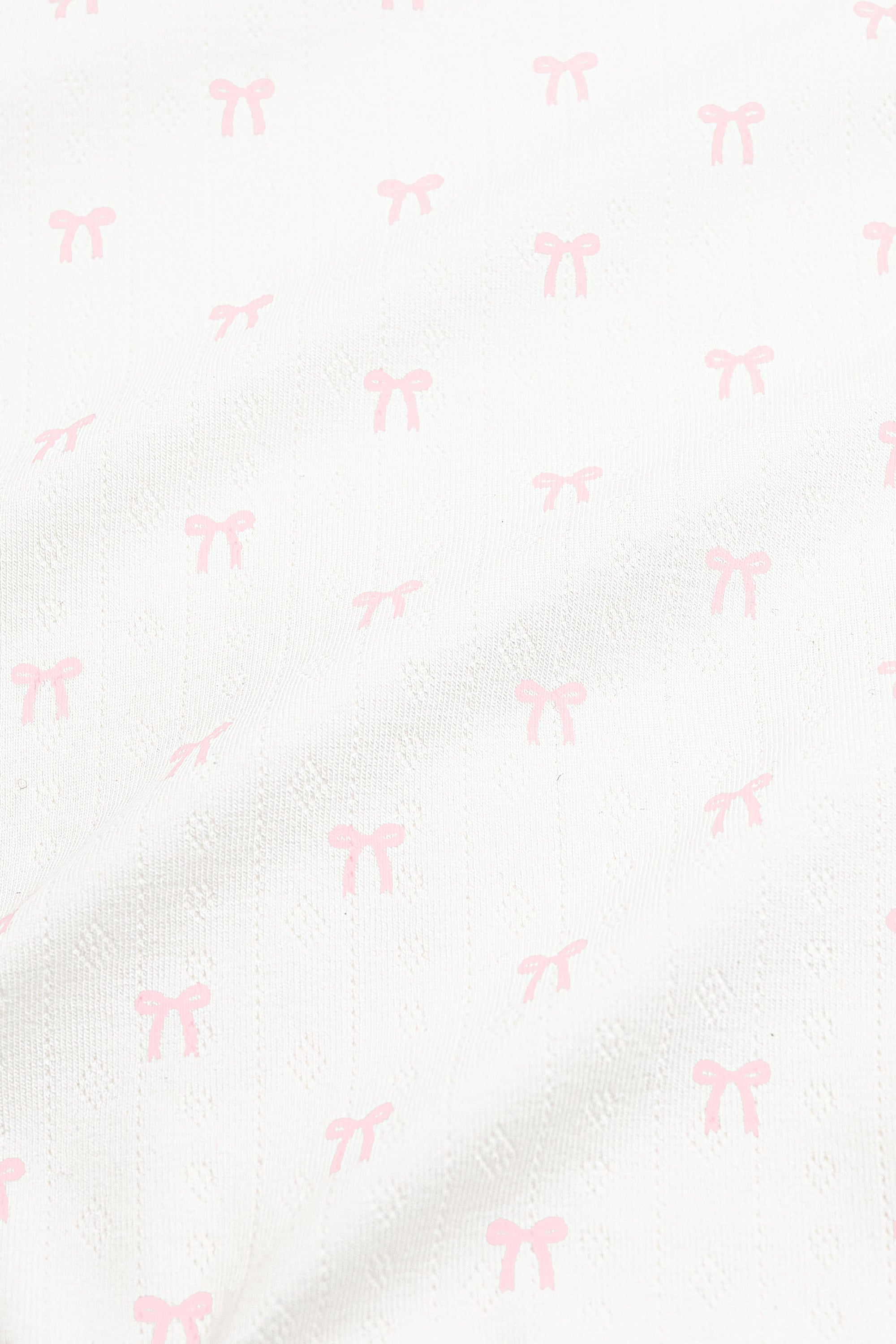Close up of the pointelle fabric: An all-over print of dainty light pink bows over a white background.