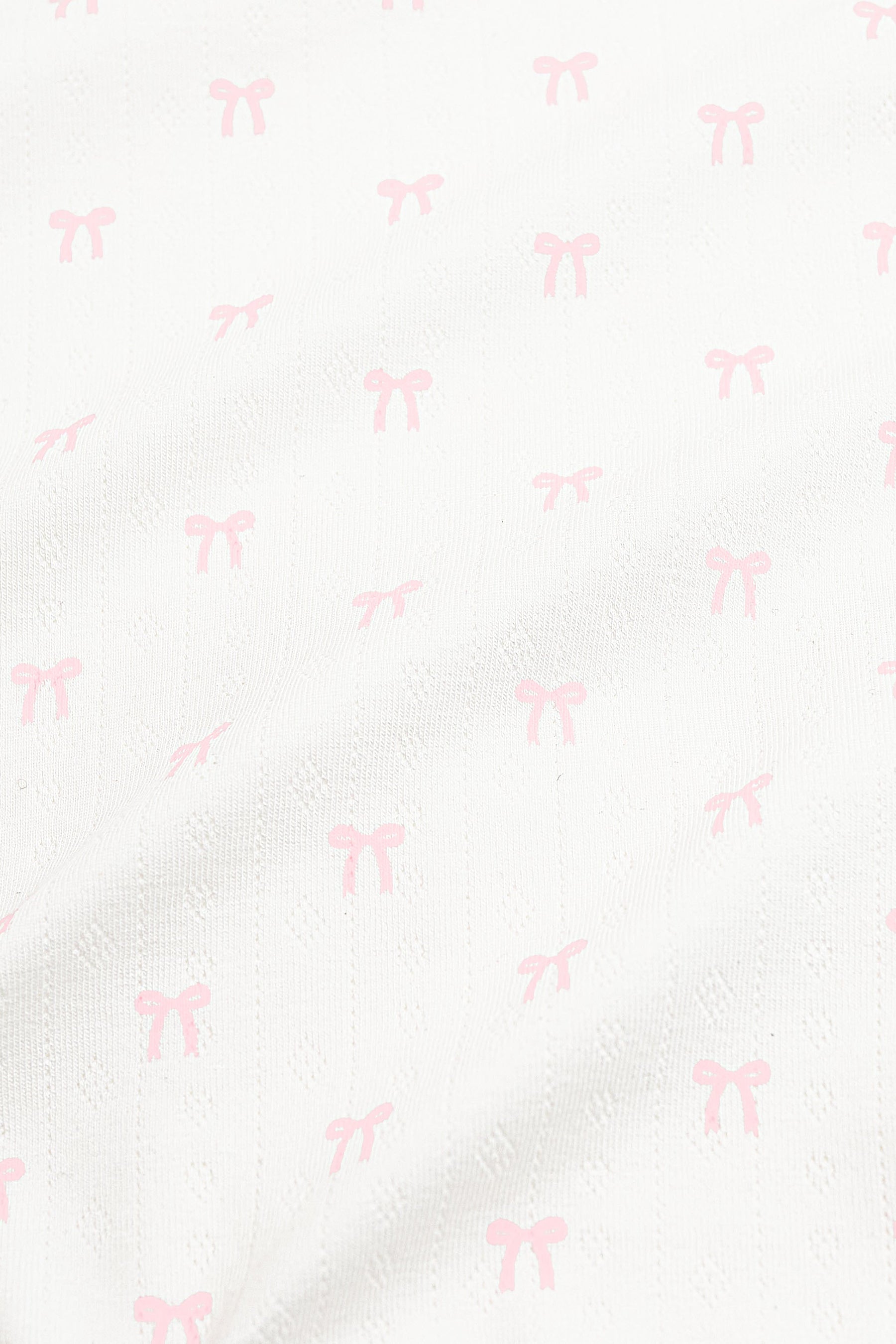 Close up of the pointelle fabric: An all-over print of dainty light pink bows over a white background.
