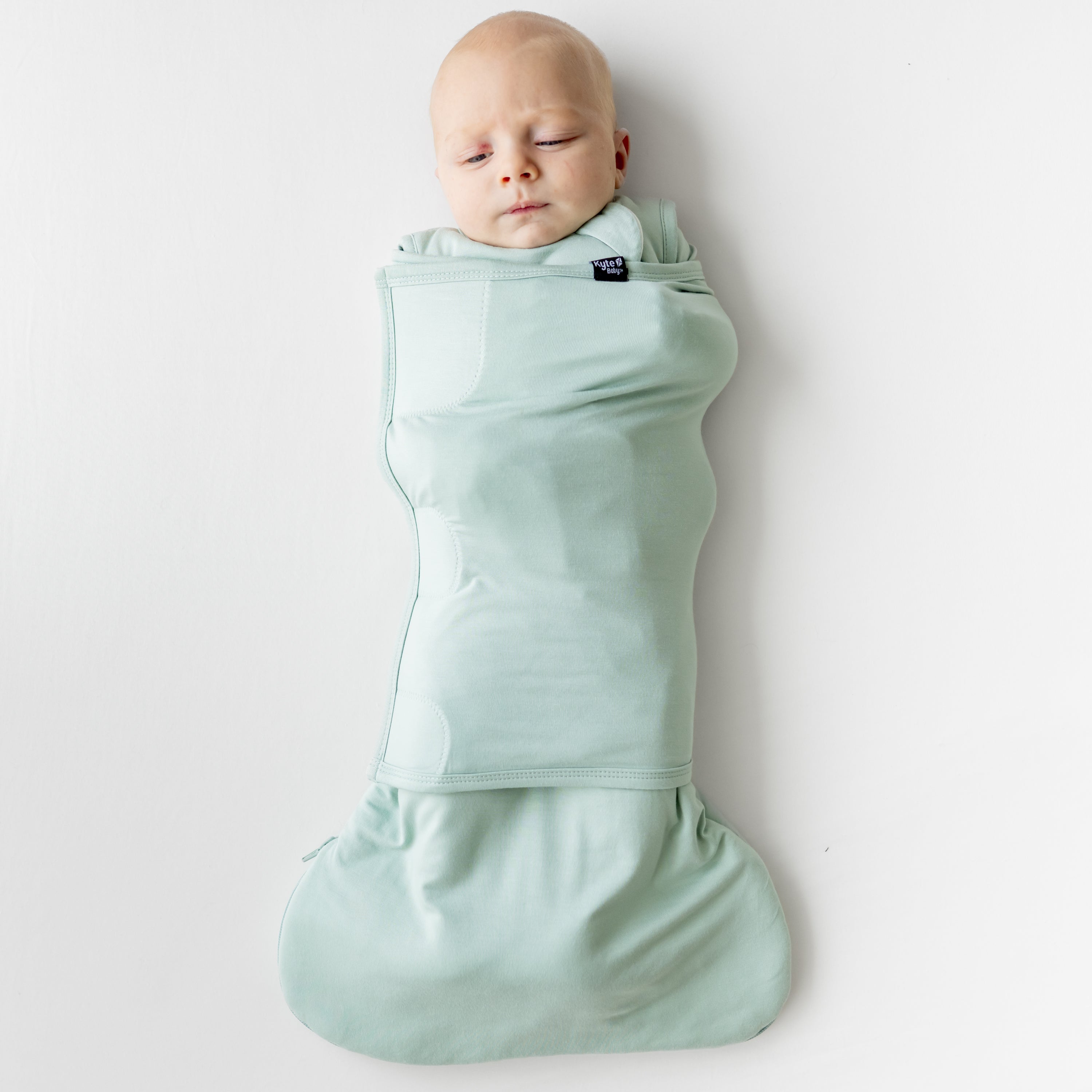 Infant laying down on a light neutral background swaddled in the Sleep Bag Swaddler in Sage 1.0