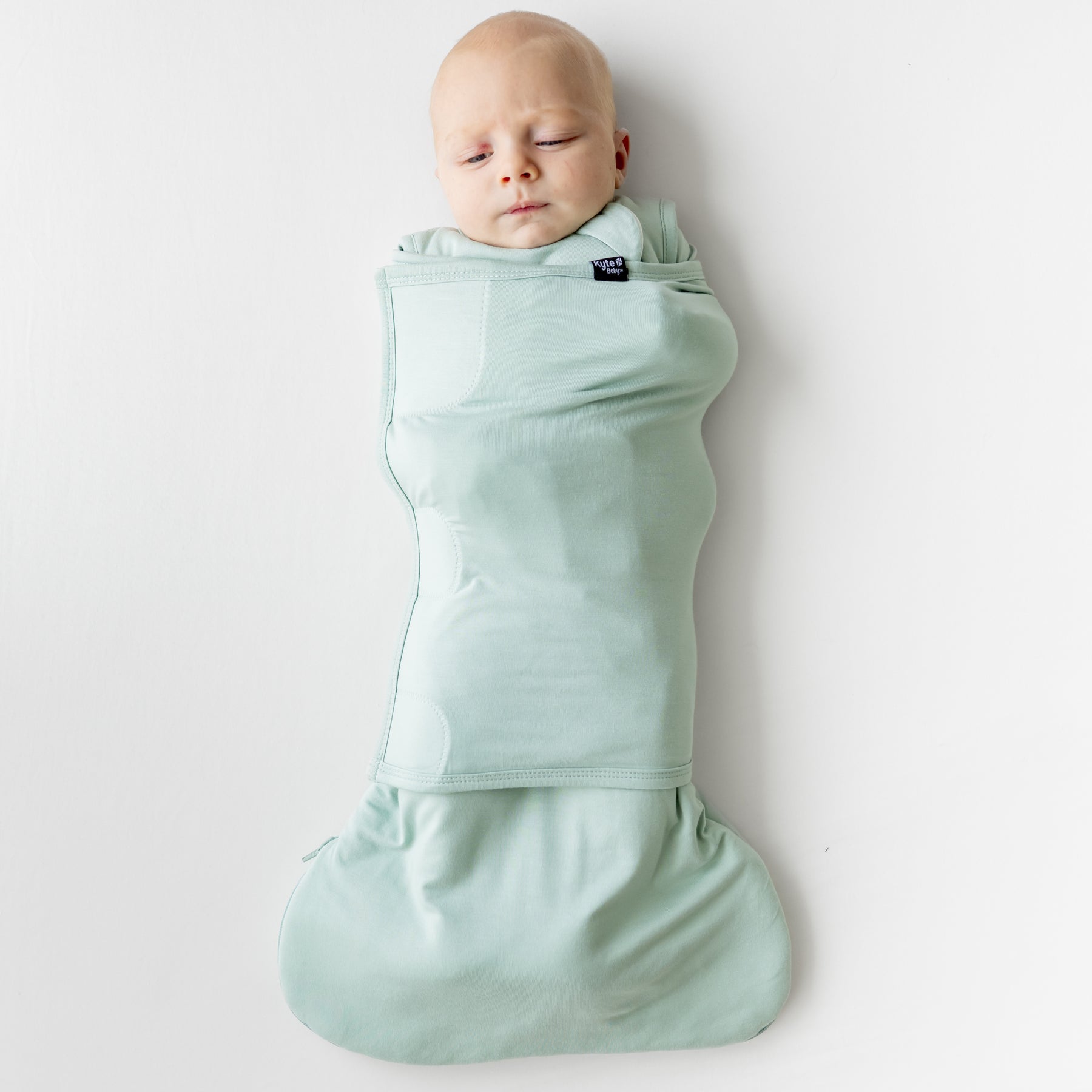 Infant laying down on a light neutral background swaddled in the Sleep Bag Swaddler in Sage 1.0