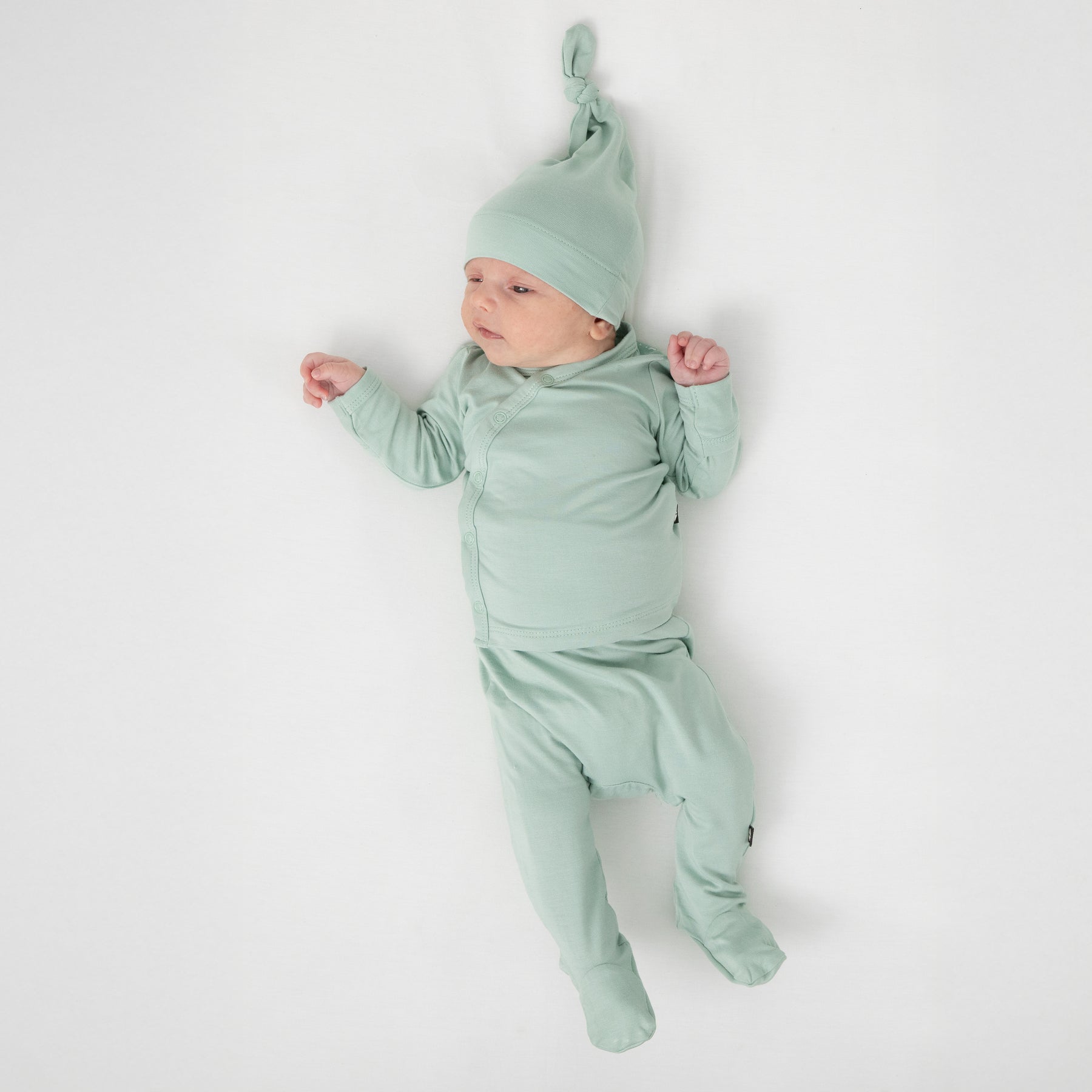 Newborn laying on a white crib sheet wearing the Take Me Home Set with Cap in Sage