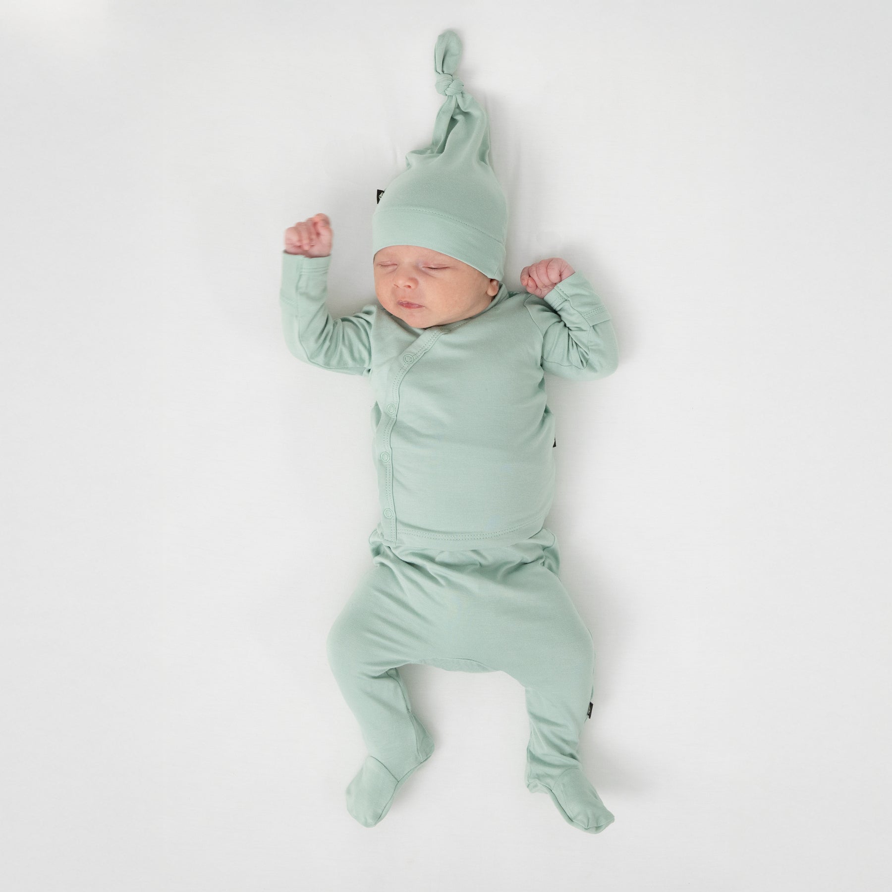 Sleeping newborn wearing the Take Me Home Set with Cap in Sage laying on a white crib sheet