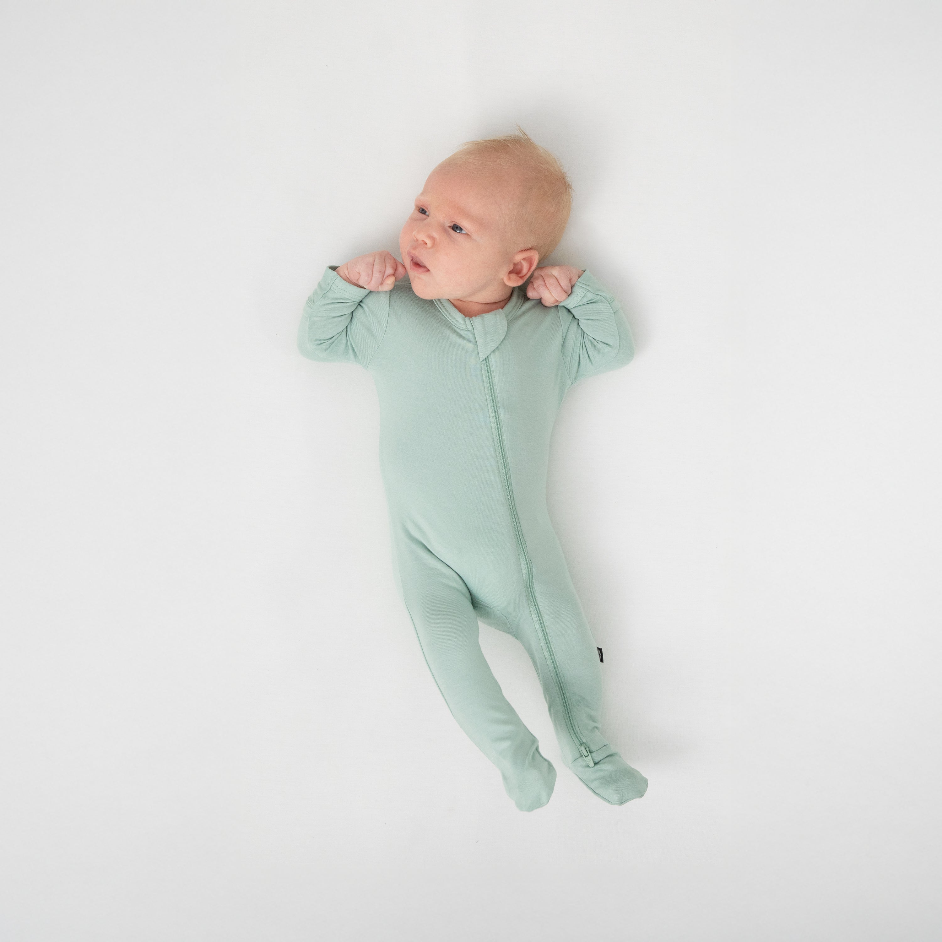 Newborn laying on a white crib sheet wearing the Zippered Footie in Sage