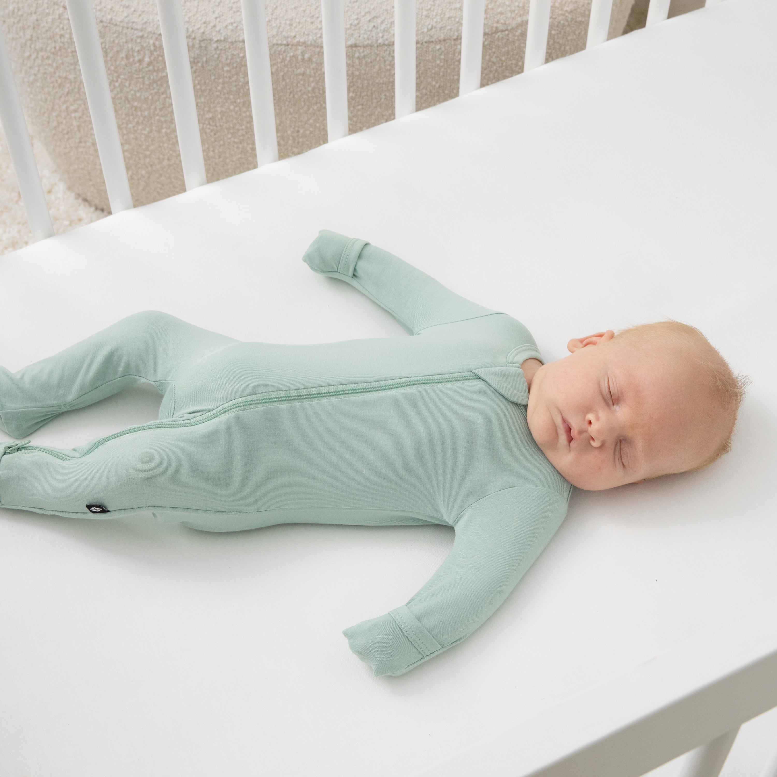 Sleeping infant laying in a crib on a white crib sheet wearing the Zippered Footie in Sage