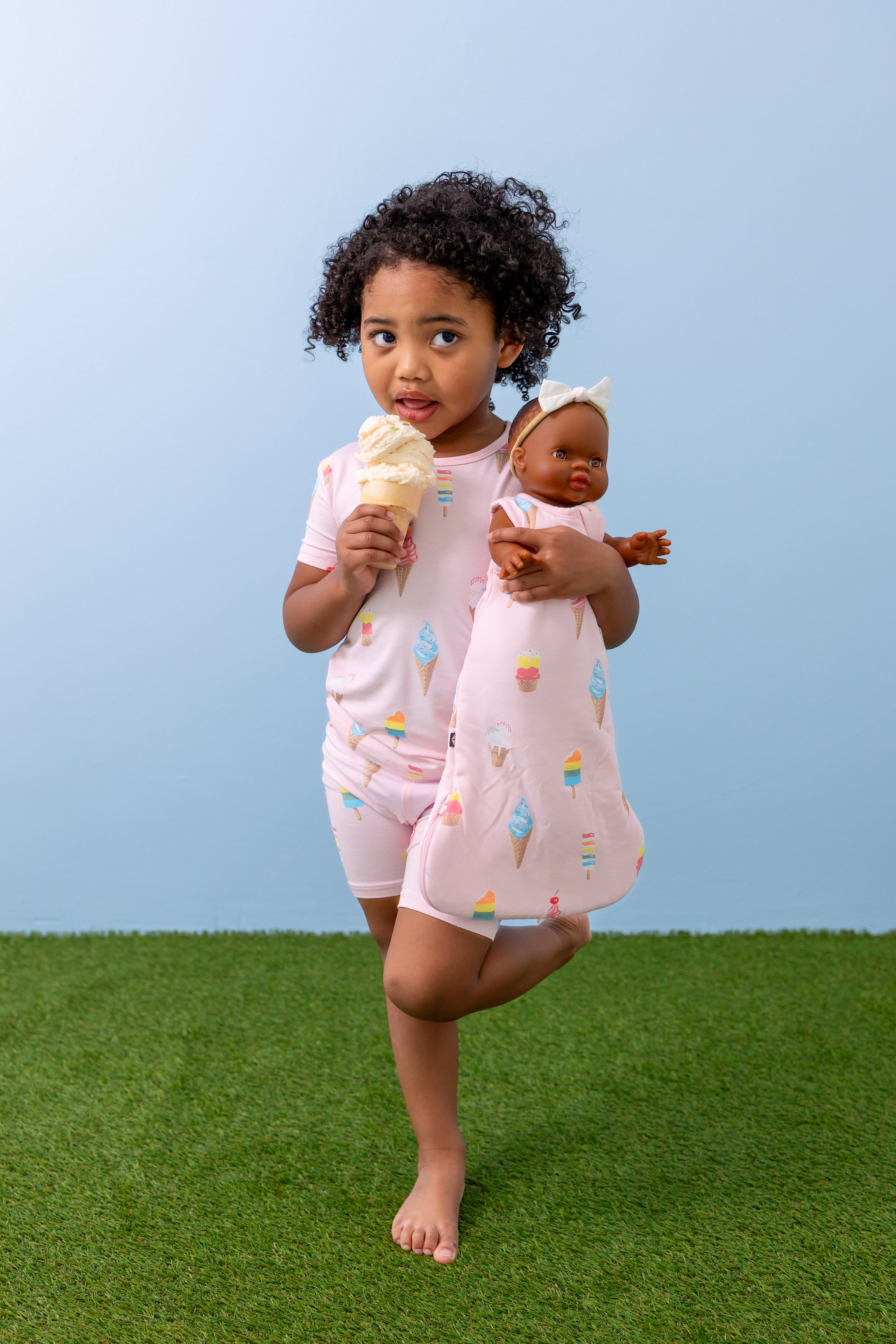 Child holding Baby doll sleep bag in Sakura Ice Cream print eating an Ice cream cone wearing a similar print Short sleeve pajama set on green grass