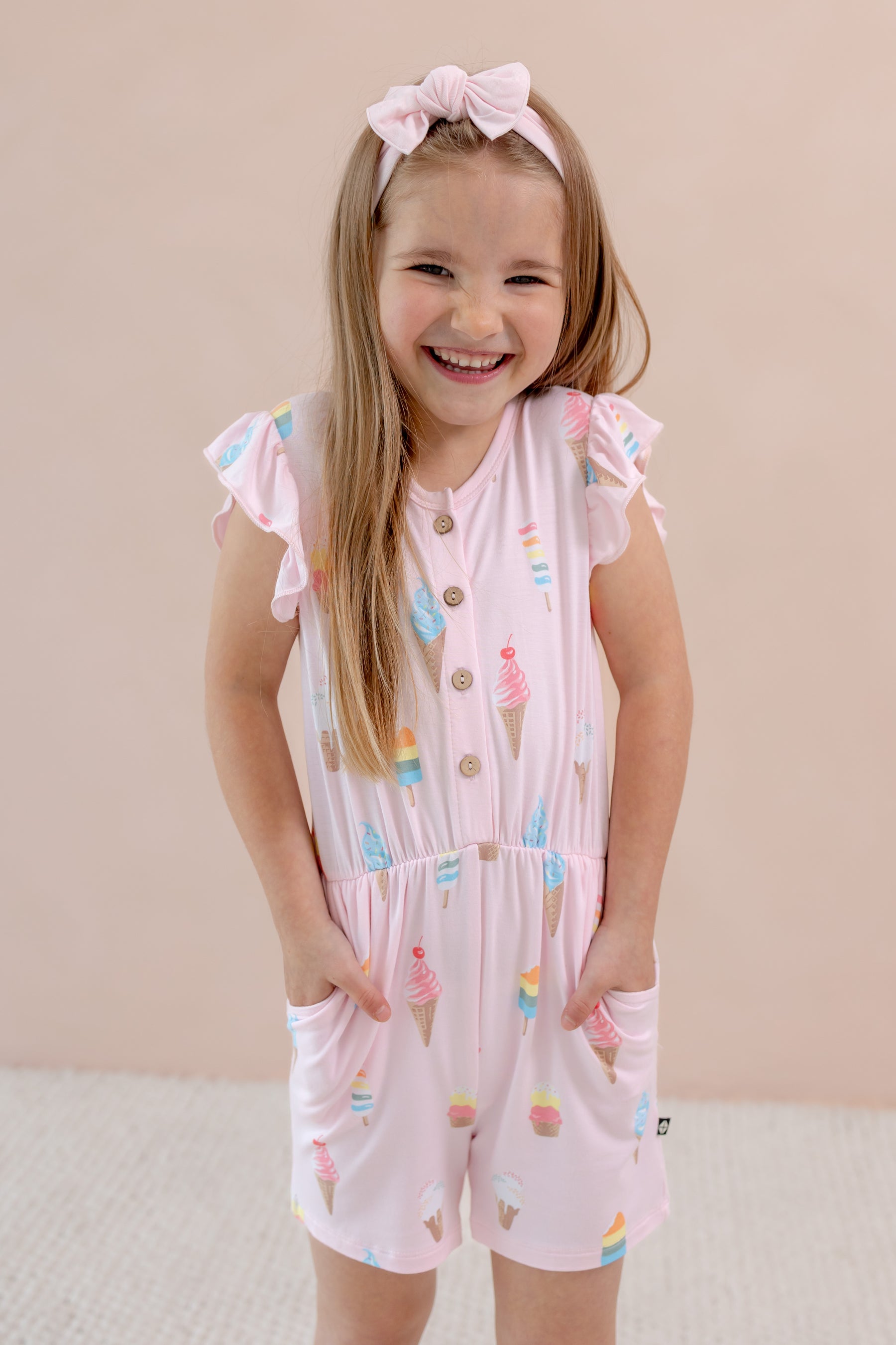 Child posing in Sakura Ice cream print Flutter Sleeve Romper with pockets on a beige background wearing a pink bow headband