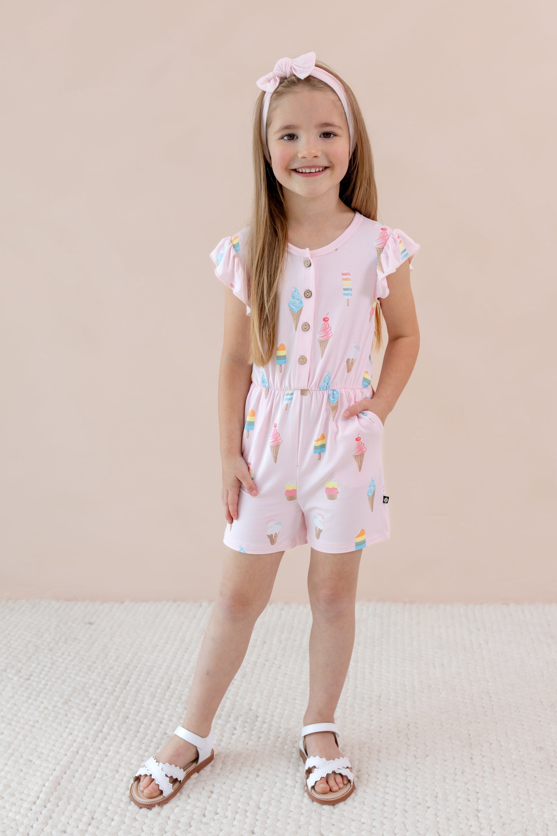 Child standing in Flutter Sleeve Romper in Sakura Ice Cream print with pink bow head band and white sandles on a beige background