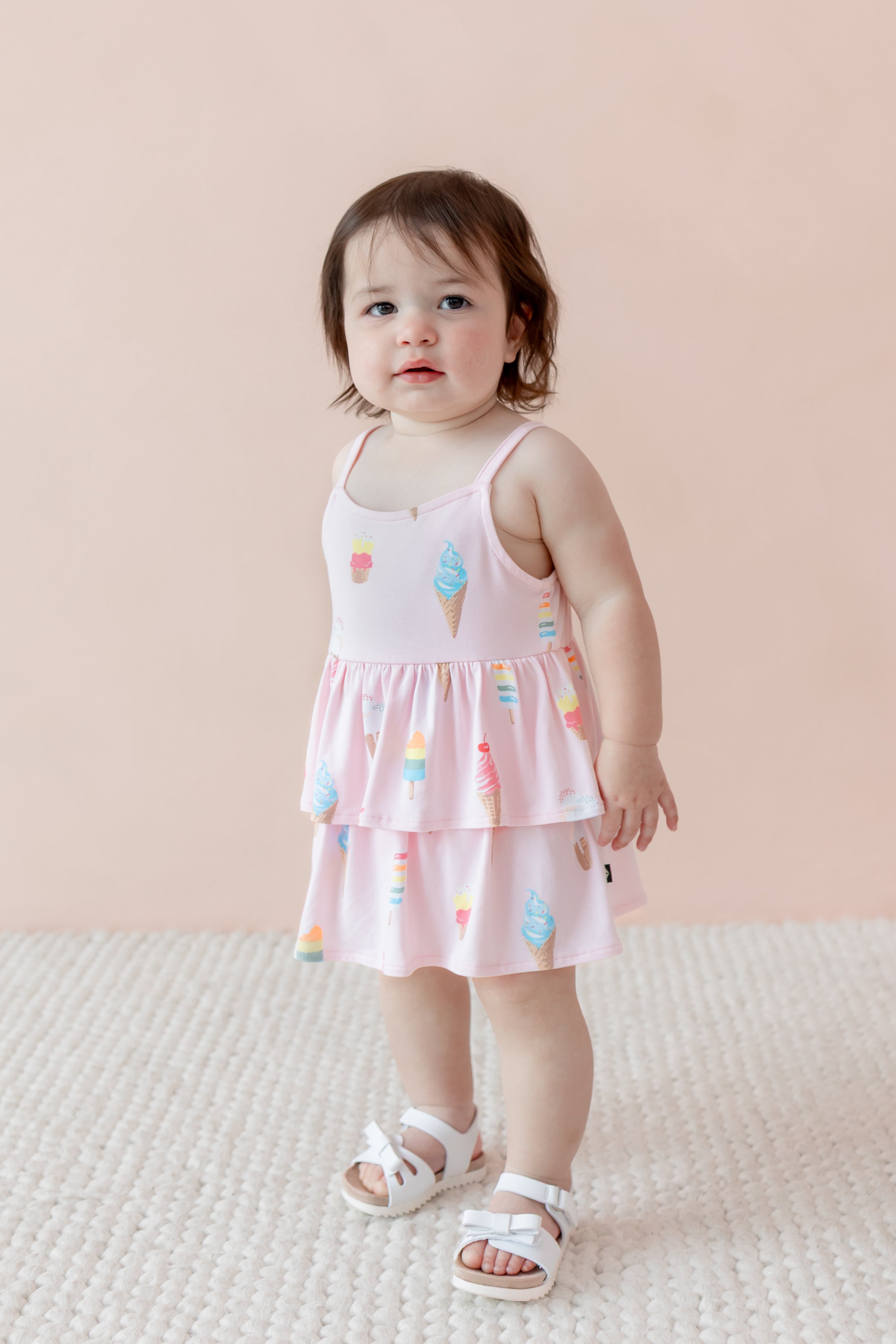 Child standing in Sakura Ice Cream print spaghetti strap ruffle romper with white sandles on beige background
