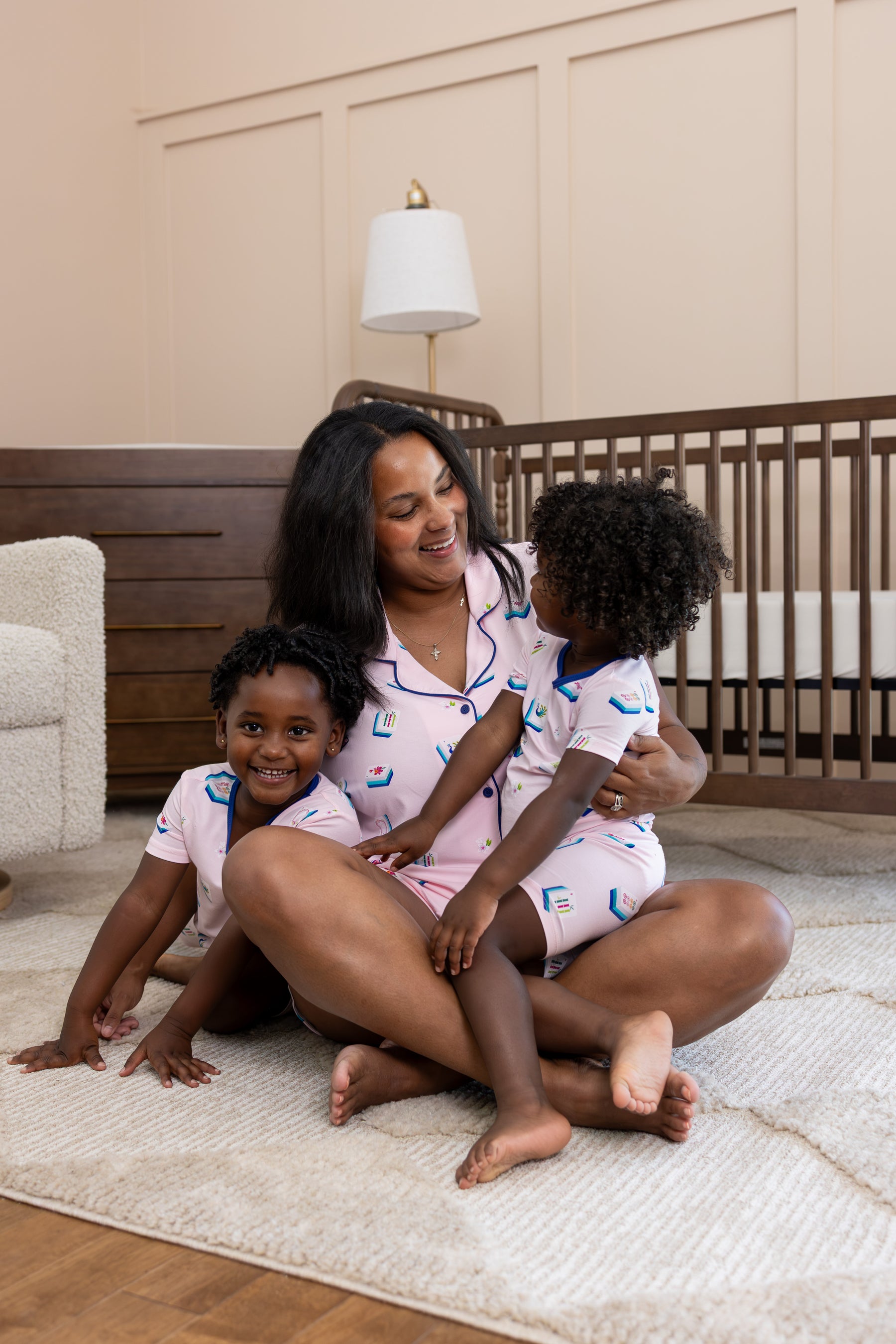 A woman and two young children wearing matching light pink pajamas with a blue mahjong pattern are sitting together on a cream rug in a nursery setting.