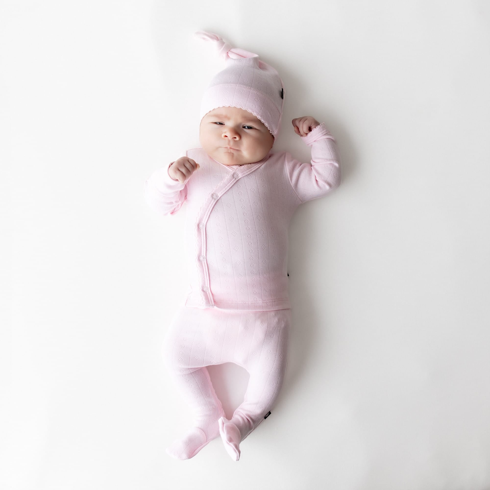 Newborn laying on a white surface wearing the Pointelle Take Me Home Set with Cap in Sakura with the picot trim visible