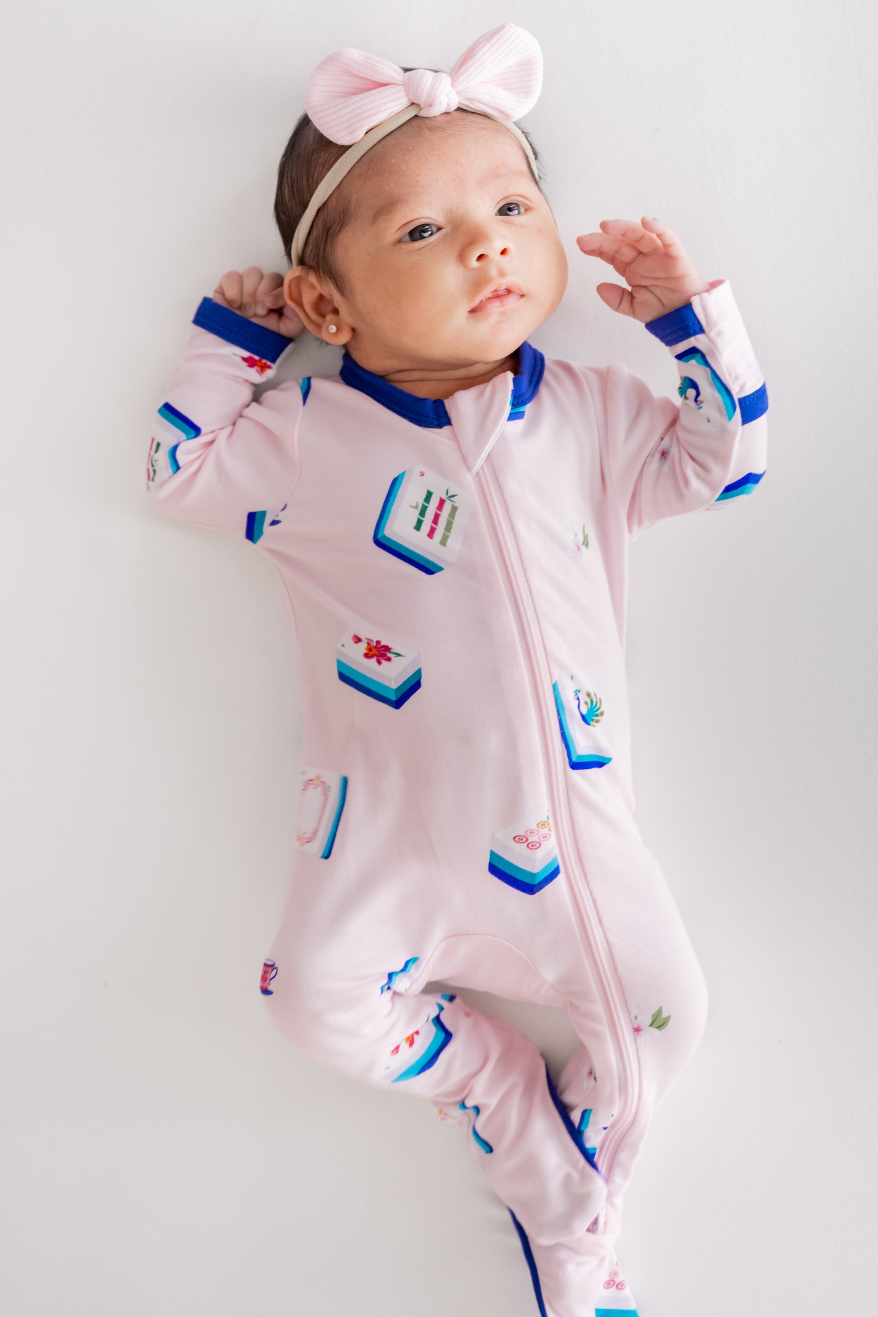 Baby wearing Zippered Footie in Sakura Mahjong print and pink bow headband lying on a beige background