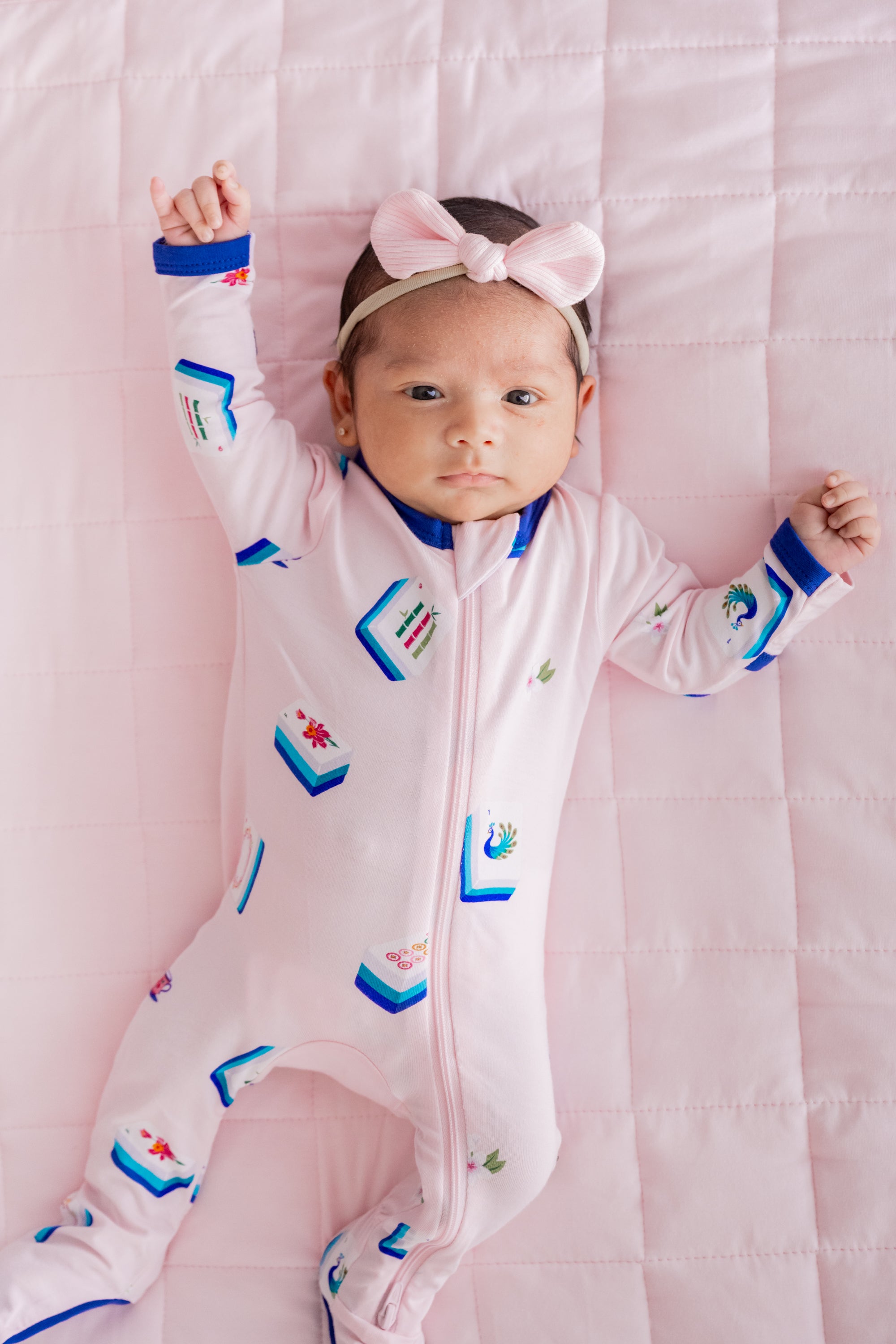 Baby wearing Zippered Footie in Sakura Mahjong print and pink bow headband lying on a pink blanket