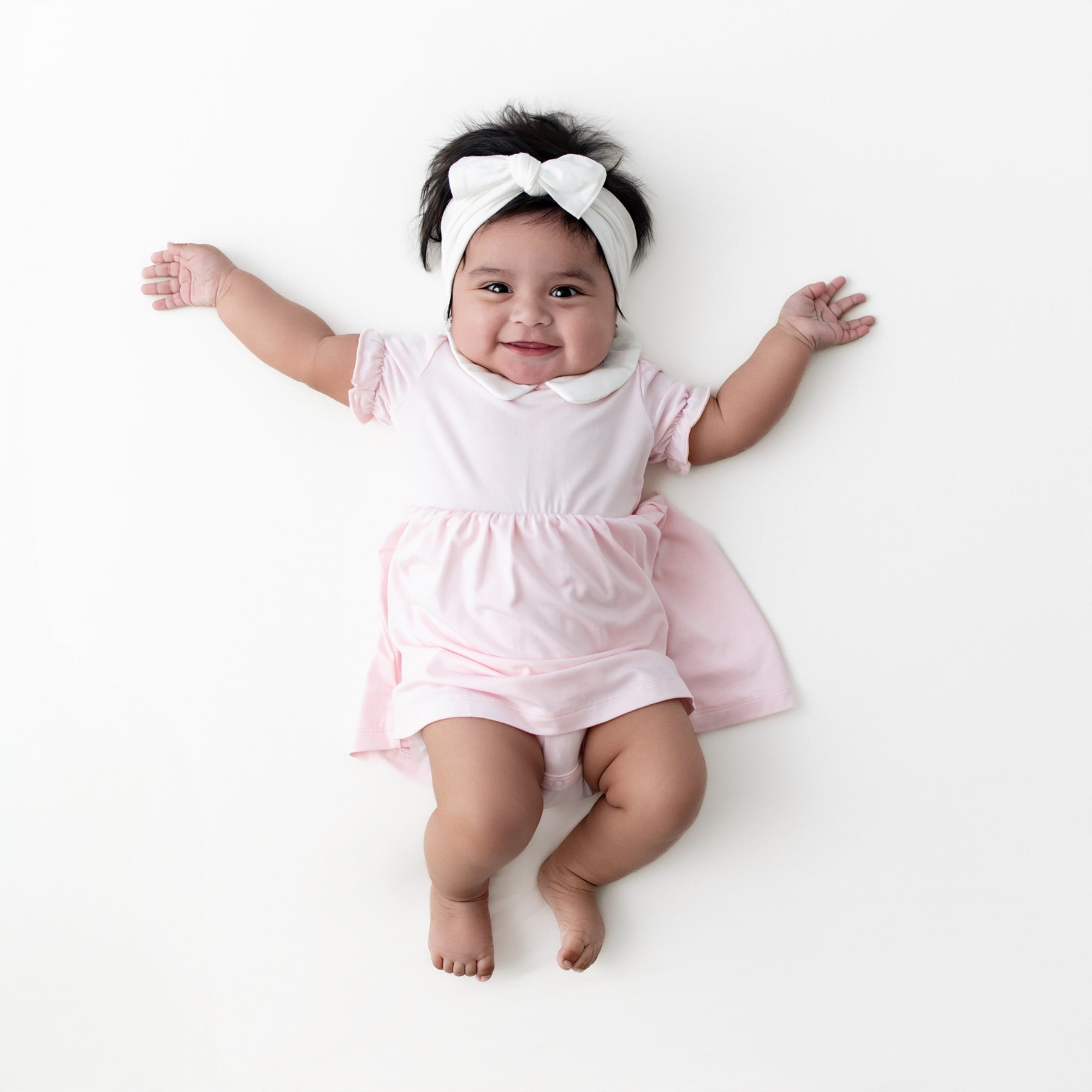 Infant girl modeling the Peter Pan Collar Bodysuit Dress in Sakura paired with a white knotted bow
