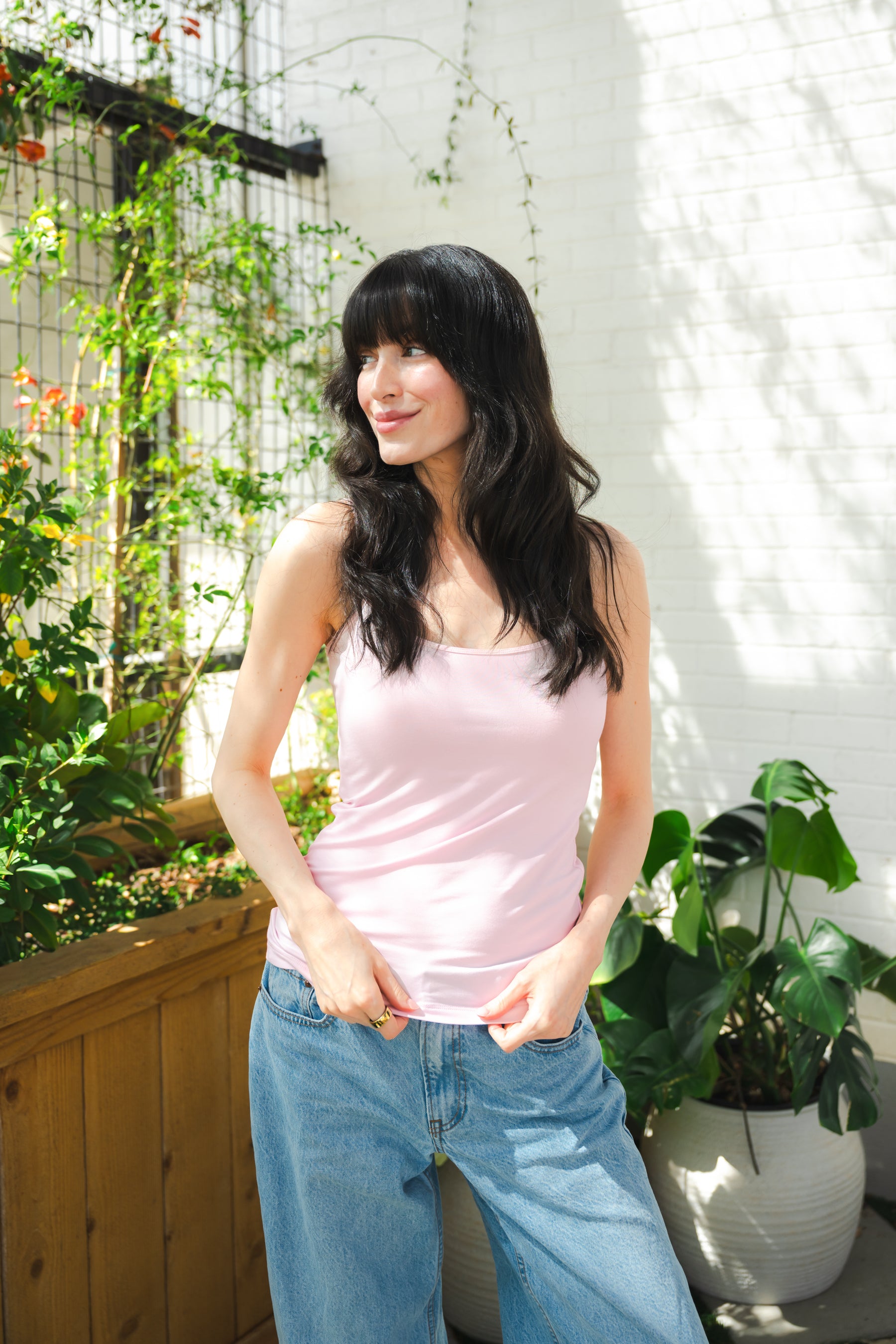 Woman wearing a pink tank top and blue jeans standing outdoors with plants in the background