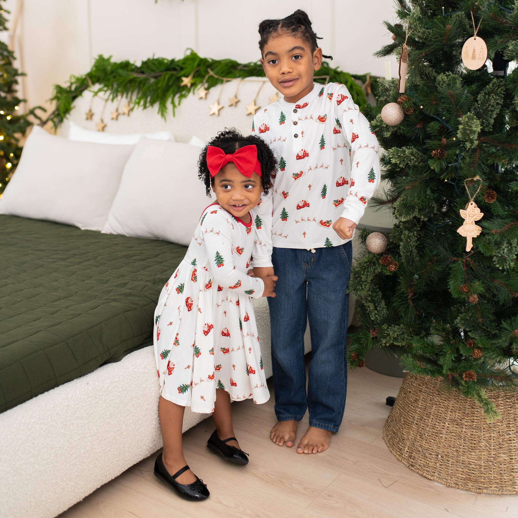 Siblings standing beside one another between a bed and christmas tree wearing the Long Sleeve Twirl Dress in Santa Sleigh and Long Sleeve Henley Tee in Santa Sleigh