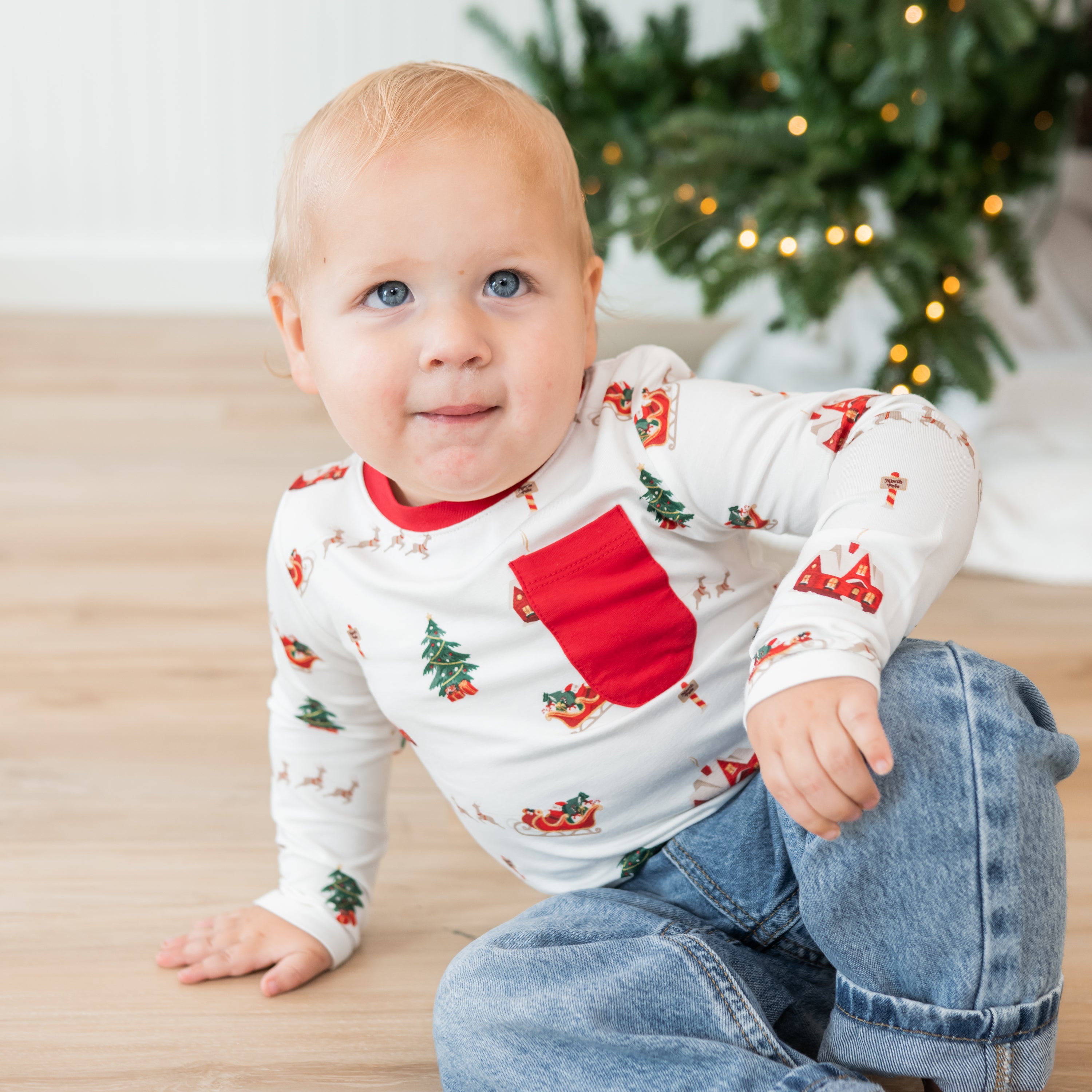 Young toddler sitting on the floor wearing the Long Sleeve Toddler Crew Neck Tee in Santa Sleigh paired with light wash jeans