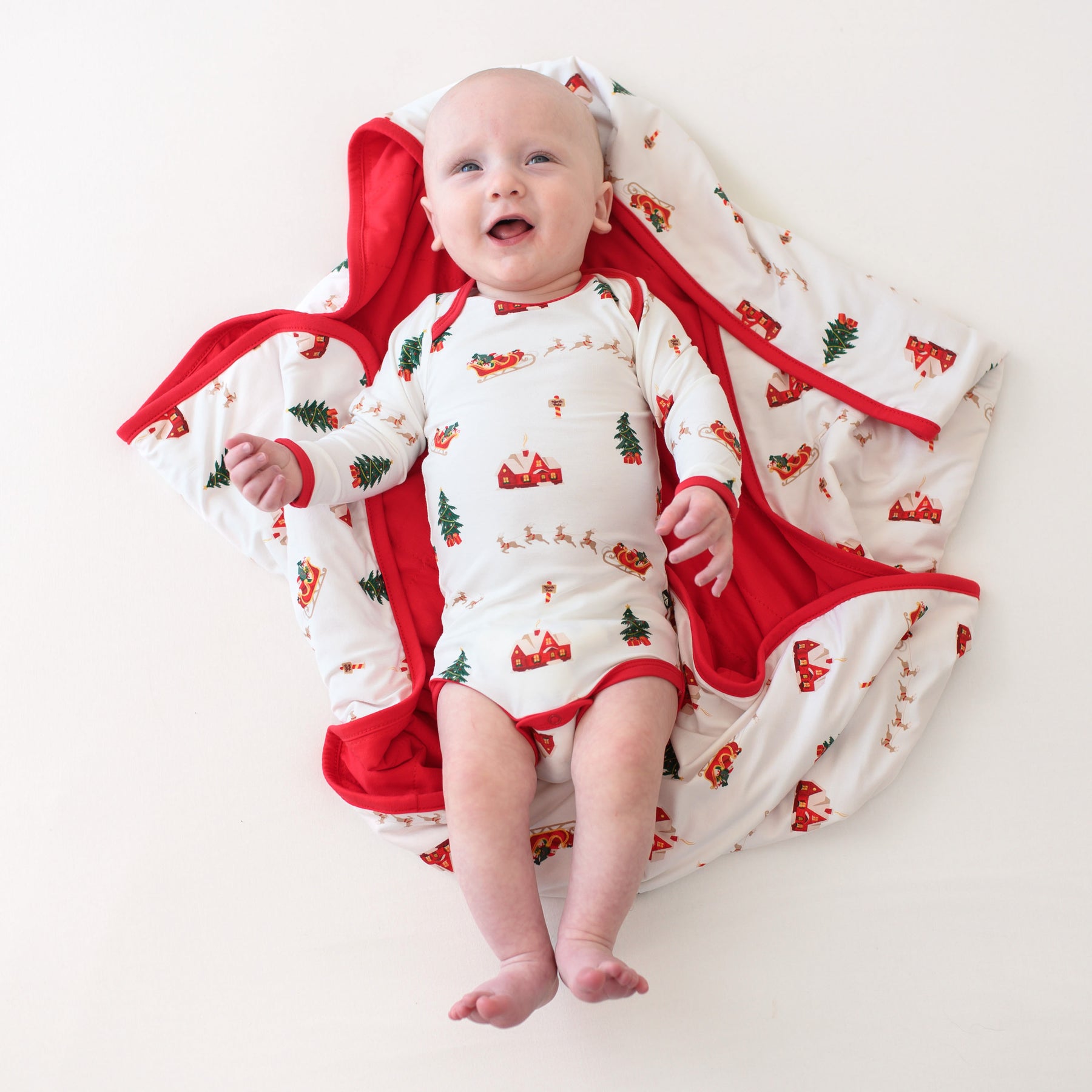 Infant laying down on a Santa Sleigh Baby blanket wearing the Long Sleeve Bodysuit in Santa Sleigh