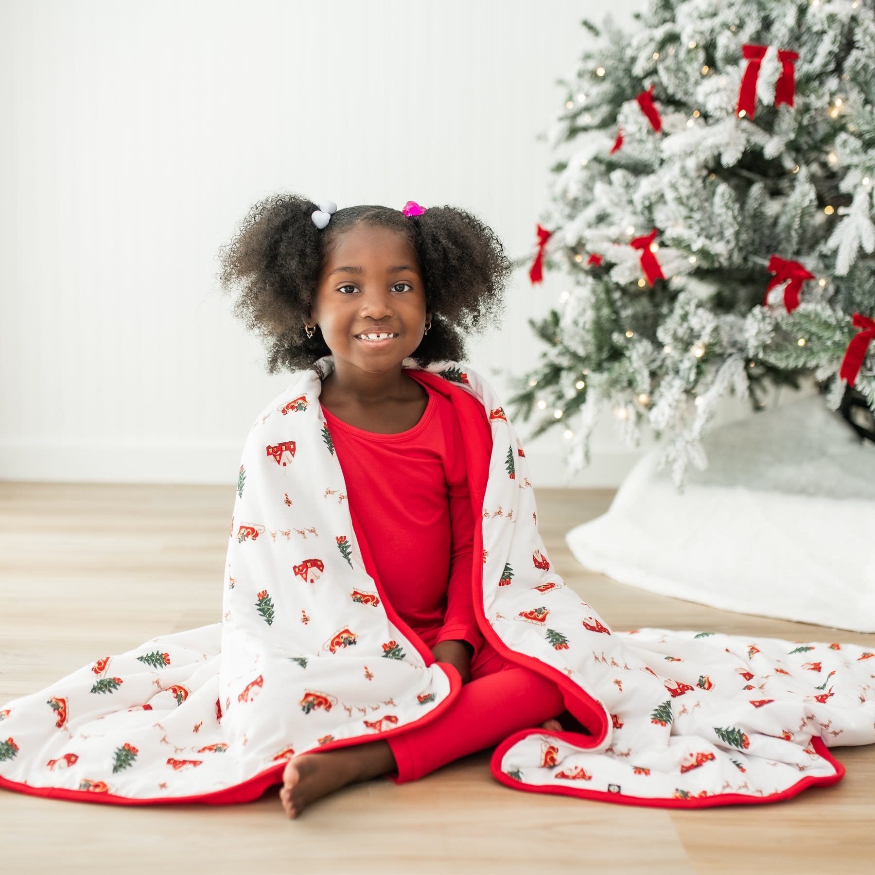 Girl sitting on the floor wearing red pajamas with the Toddler Blanket in Santa Sleigh 1.0 over her shoulders