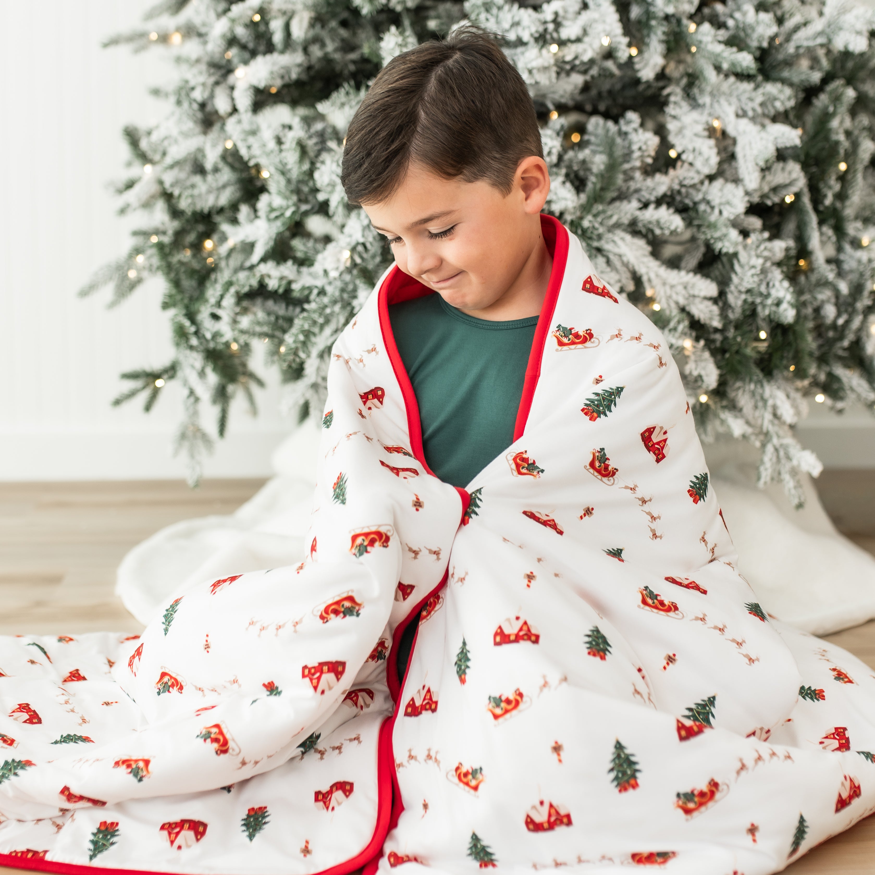 Young boy sitting on the floor wearing green pajamas with the Youth Blanket in Santa Sleigh 2.5 over his shoulders
