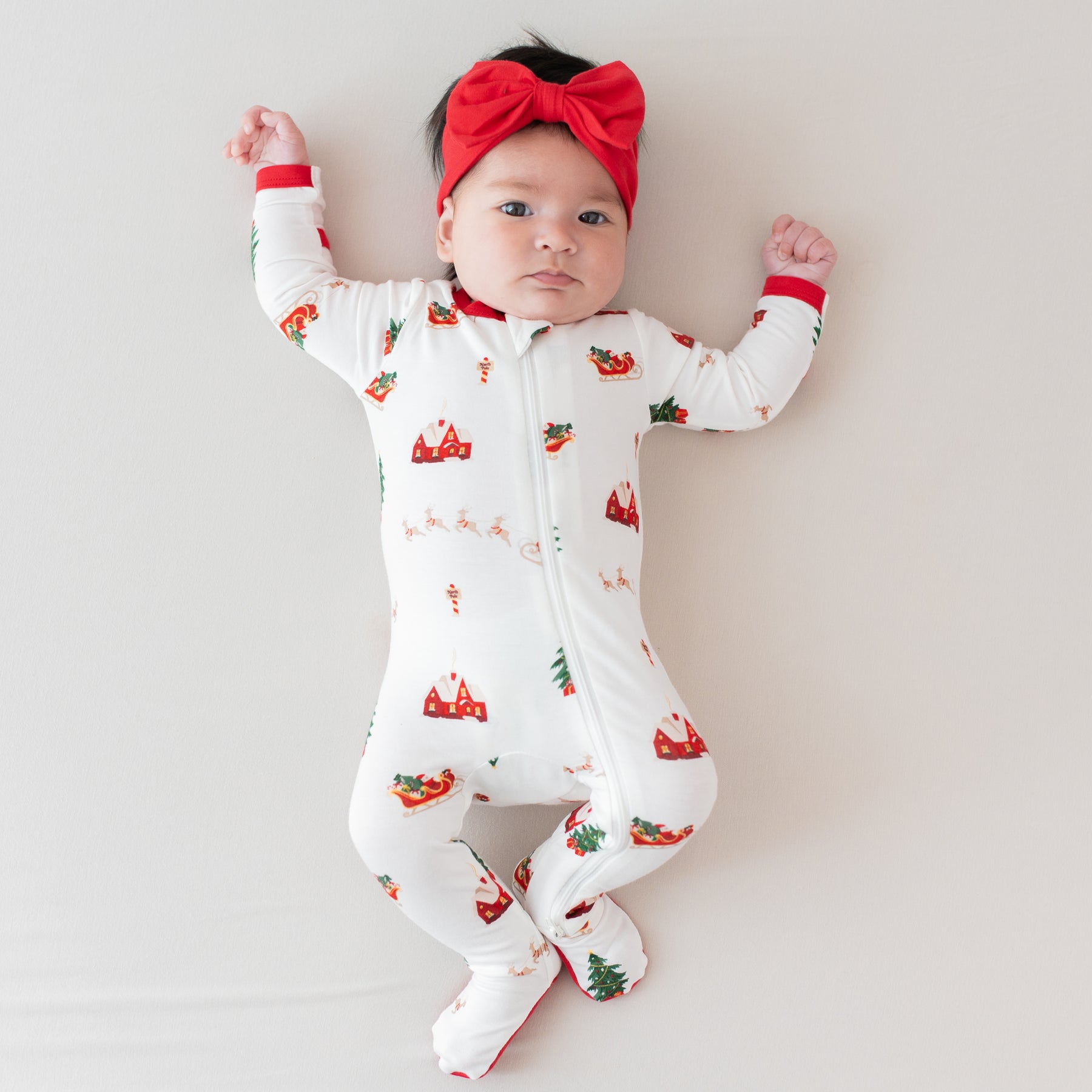 Infant girl laying down wearing the Zippered Footie in Santa Sleigh paired with a bow headband in cardinal