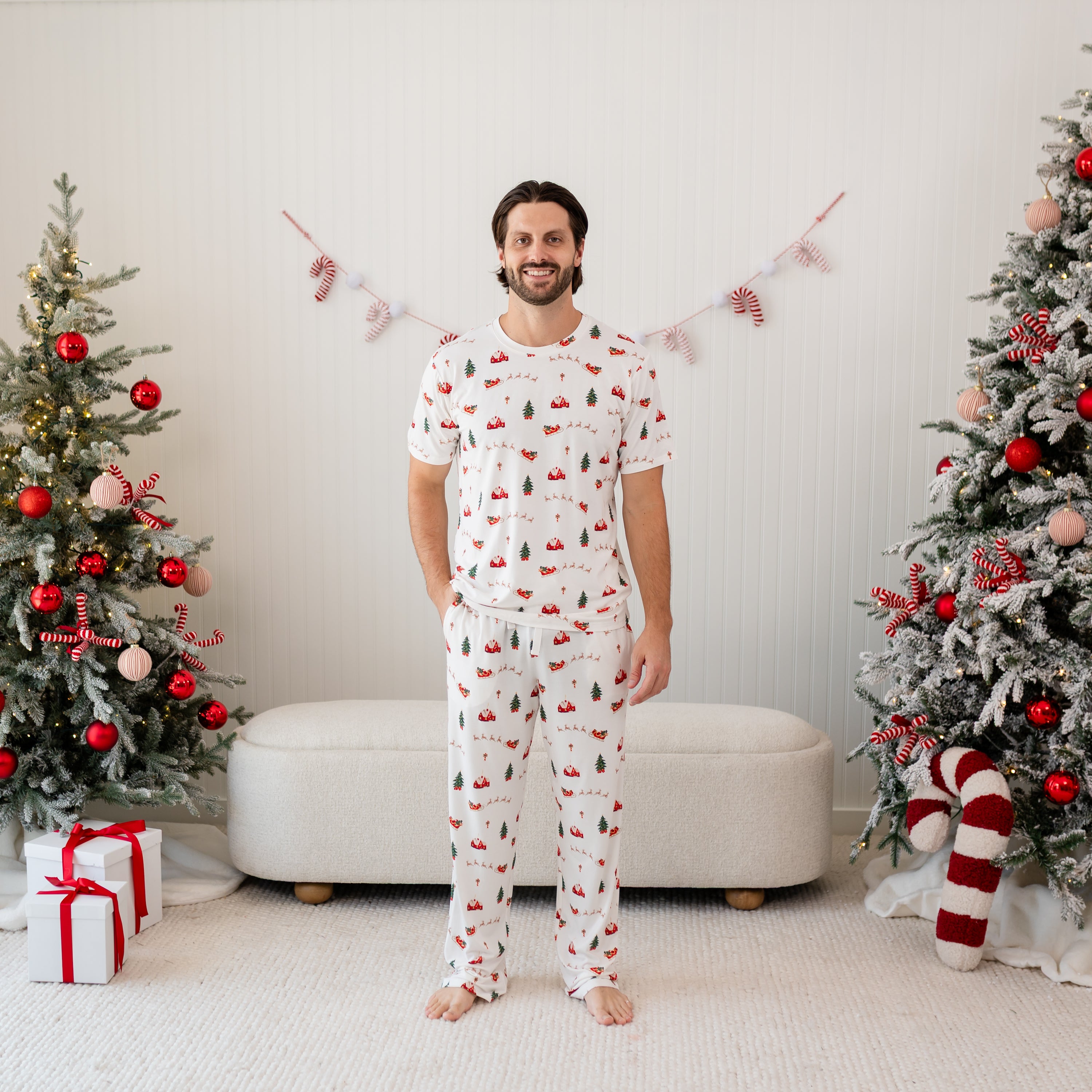 Male model standing in front of a christmas inspired back drop of Christmas trees and presents wearing the Men's Crew Neck Tee in Santa Sleigh and matching men's lounge pant