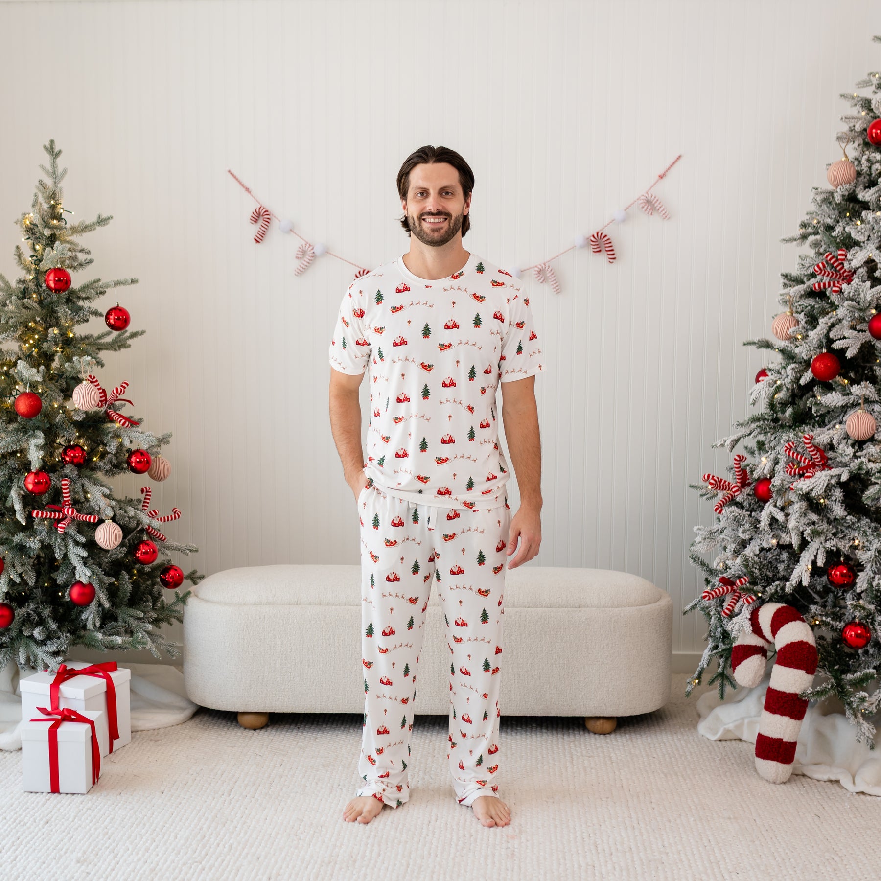 Male model standing in front of a christmas inspired back drop of Christmas trees and presents wearing the Men's Crew Neck Tee in Santa Sleigh and matching men's lounge pant