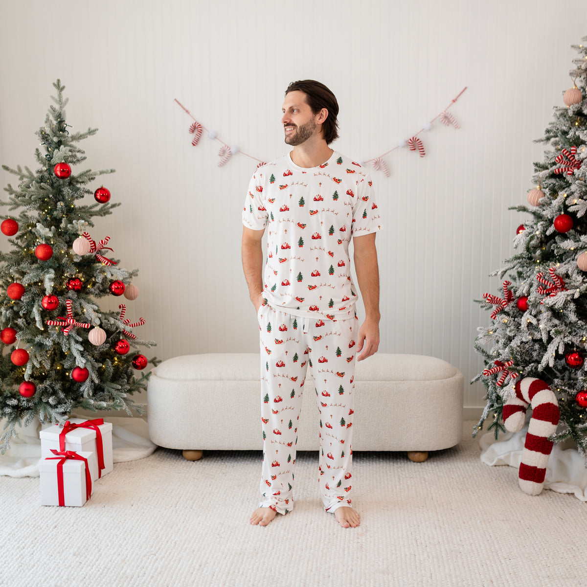 Male model standing between two christmas tree's wearing the Men's Lounge Pants in Santa Sleigh and matching Men's Crew Neck tee