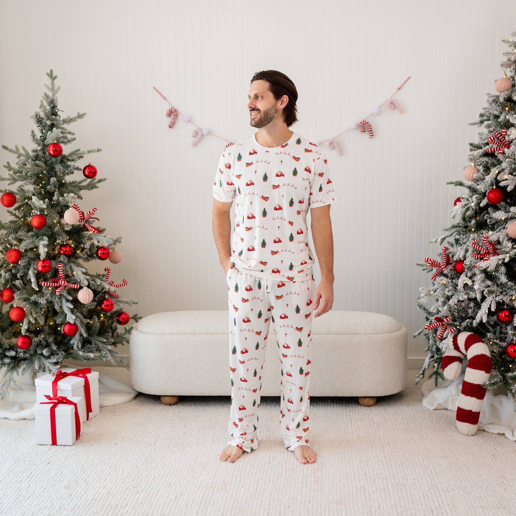 Male model standing between two christmas tree's wearing the Men's Lounge Pants in Santa Sleigh and matching Men's Crew Neck tee