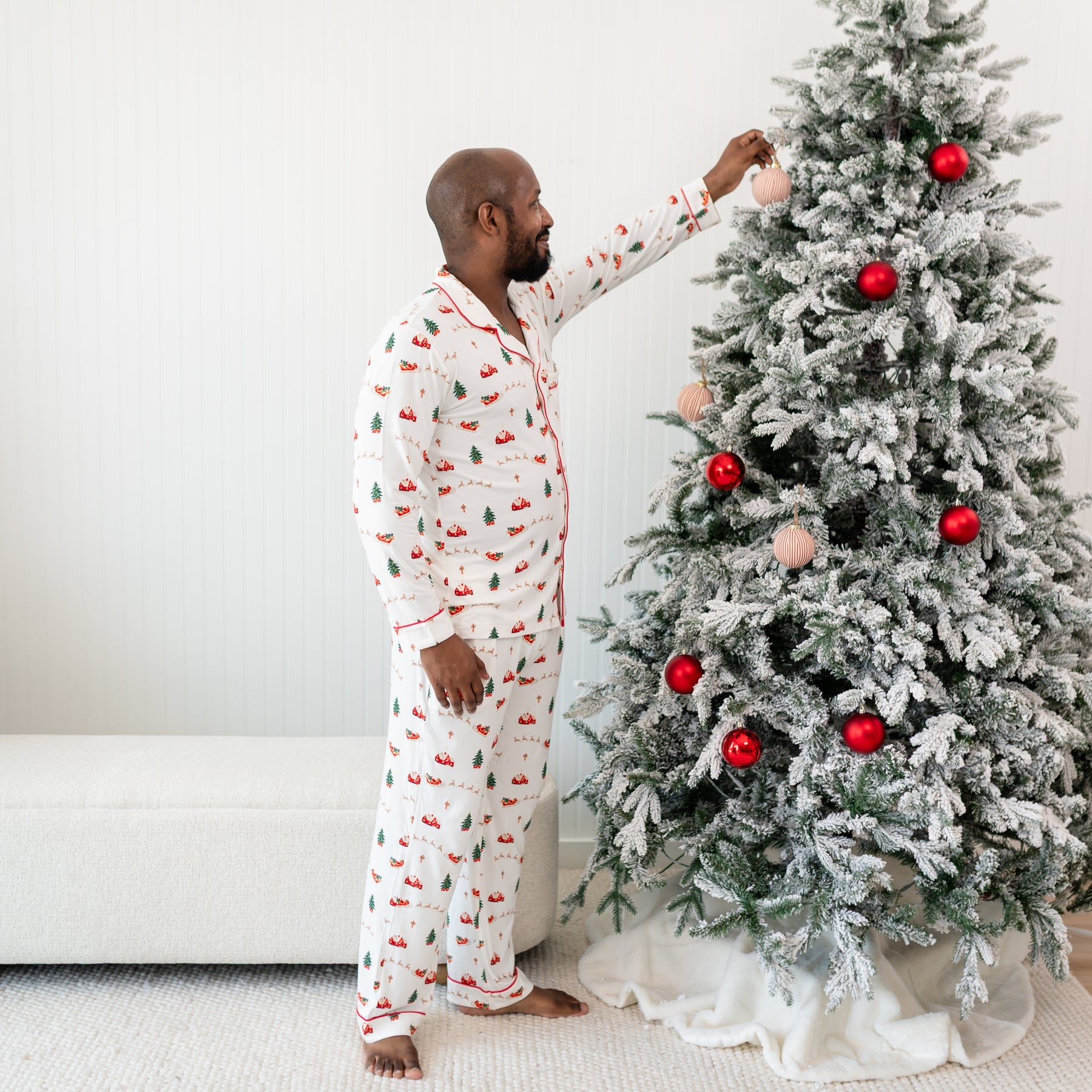 Male Model putting an ornament on a Christmas tree wearing the Men's Long Sleeve Pajama Set in Santa Sleigh