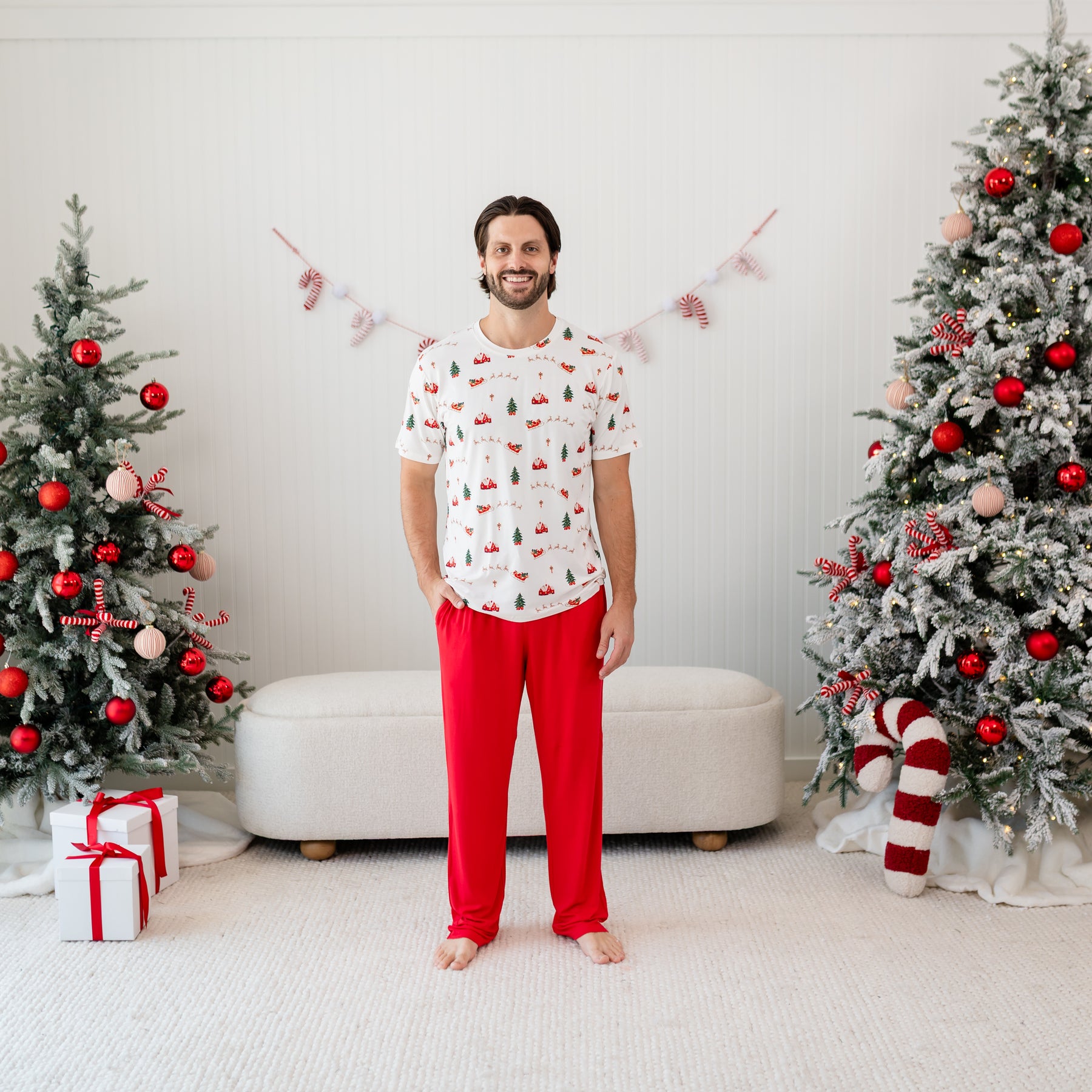 Male standing in front of a cream ottoman and between two decorated Christmas trees wearing the Men's Lounge Pants in Cardinal paired with a Men's crew neck in Santa Sleigh