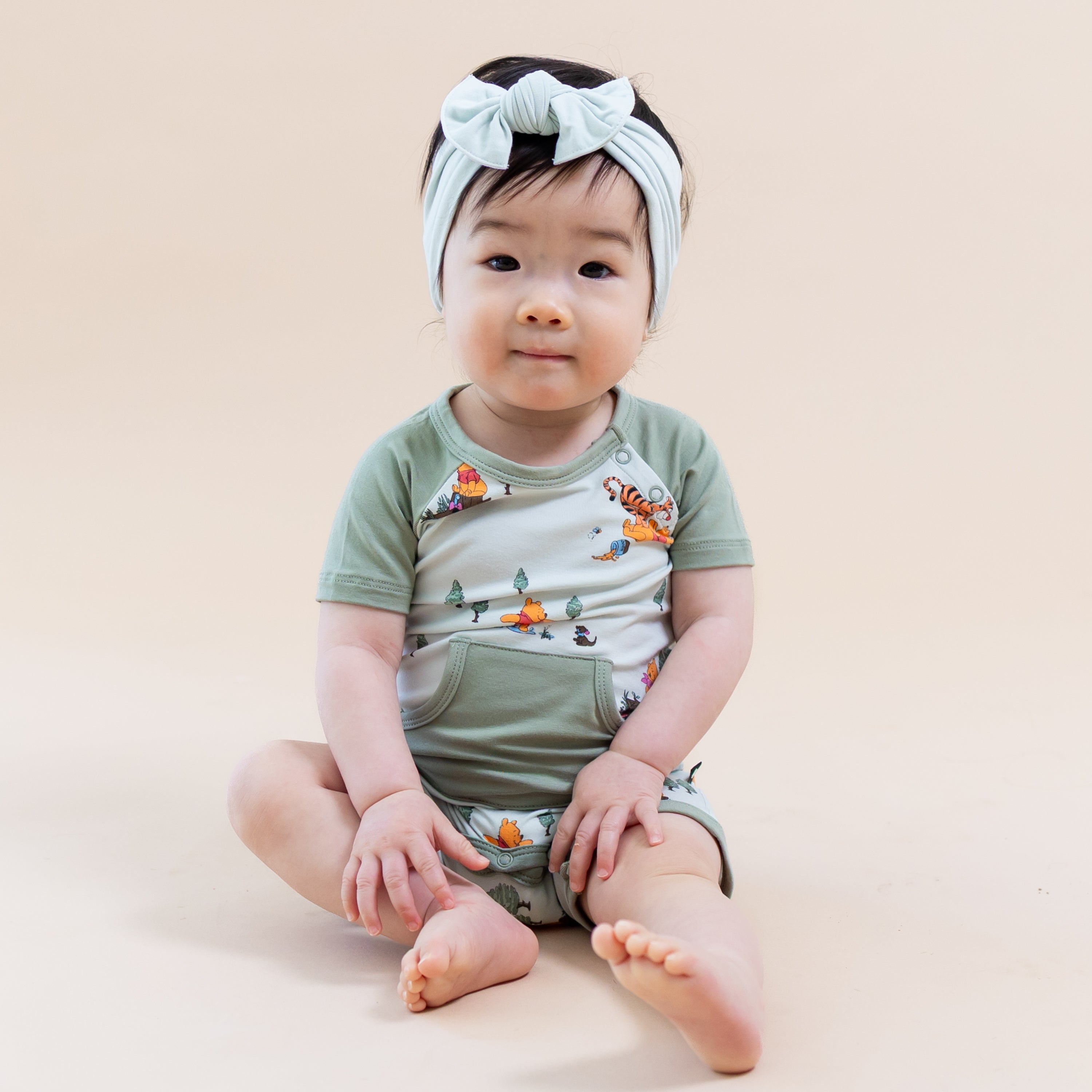 Young infant girl sitting on the floor wearing the Shortall in Winnie the Pooh Hundred Acre Wood paired with a light green bow