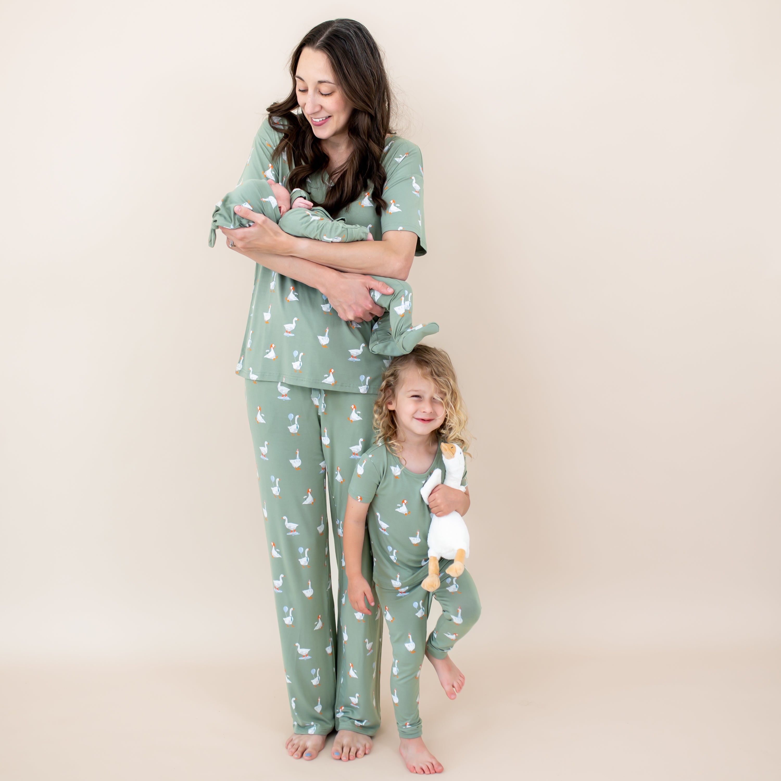 Family matching in Silly Goose. Mother wearing the Women's Lounge Pants in Silly Goose and women's v-neck holding her newborn baby wearing the take me home set and standing beside her oldest child who is in the short sleeve with pants pajama set holding a stuffed toy goose
