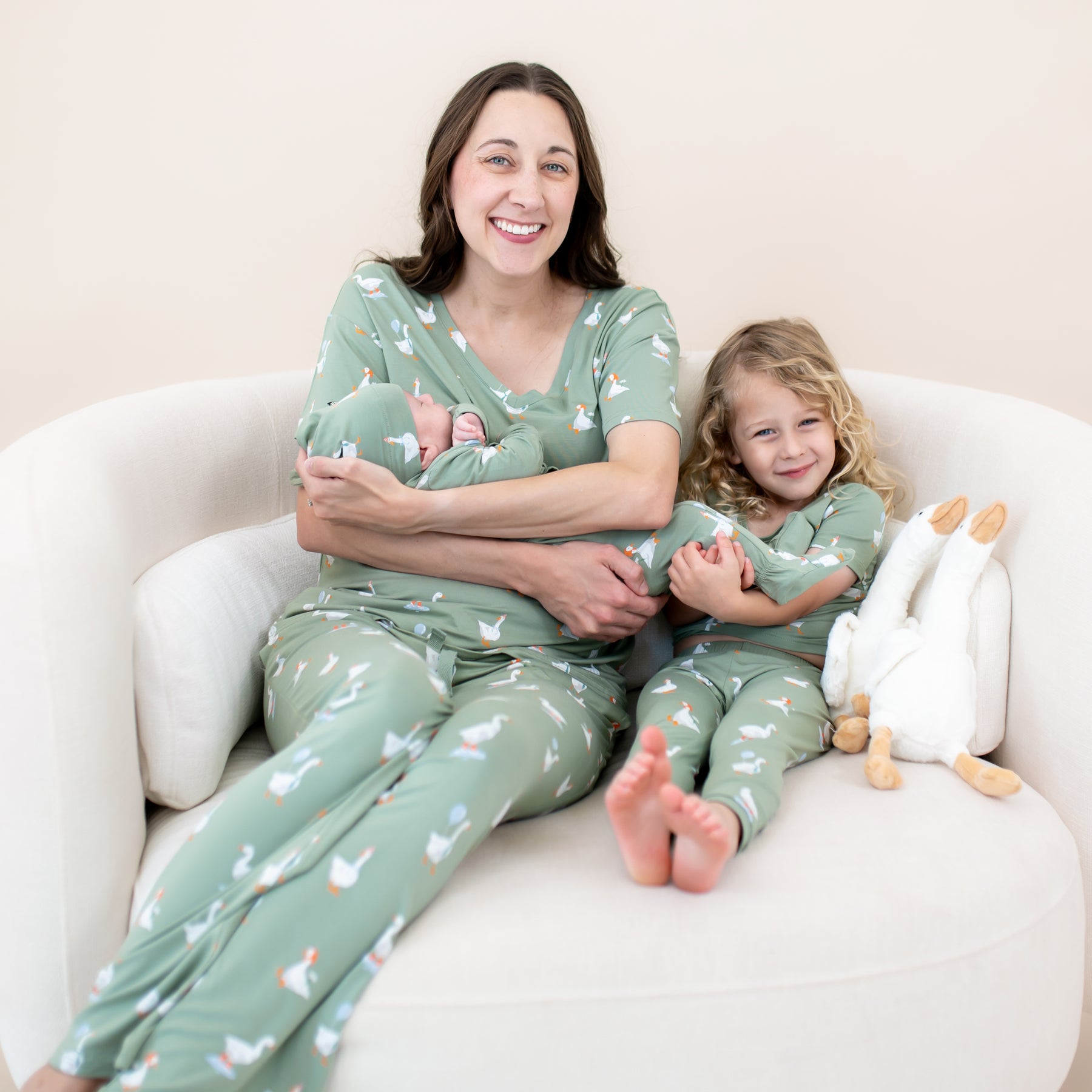 Family sitting on a cream round sofa matching in Silly Goose. Mother wearing the Women's Lounge Pants in Silly Goose and women's v-neck holding her infant wearing the take me home set sitting beside her oldest in the short sleeve with pants pajama set