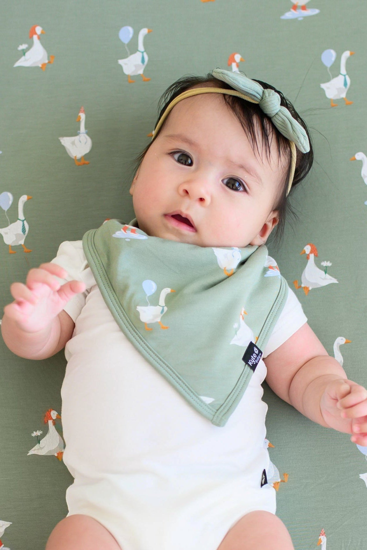 Infant laying on a Silly goose crib sheet wearing the Bib in Silly Goose, cloud bodysuit and ribbed nylon bow in Thyme