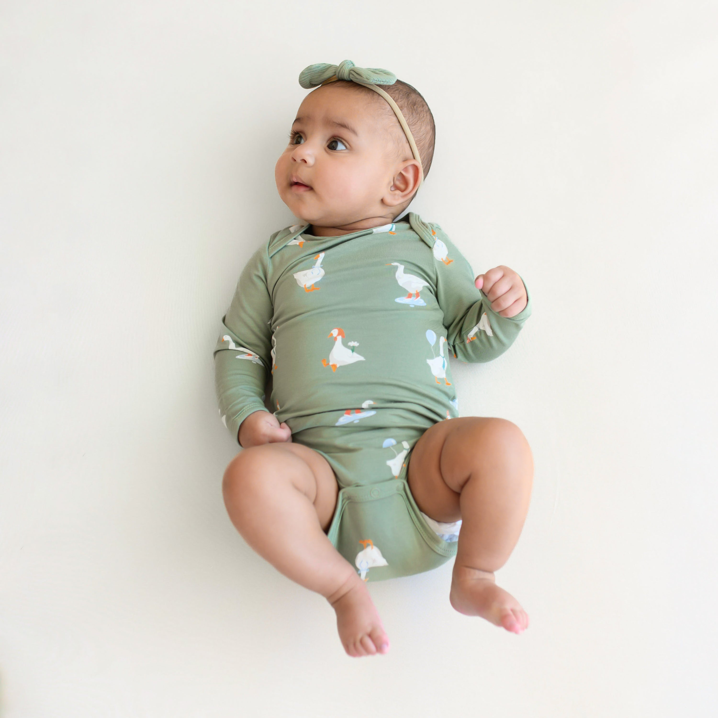Infant wearing the Long Sleeve Bodysuit in Silly Goose and Ribbed Nylon Tiny Bow in Thyme looking off to the side with her legs in the air
