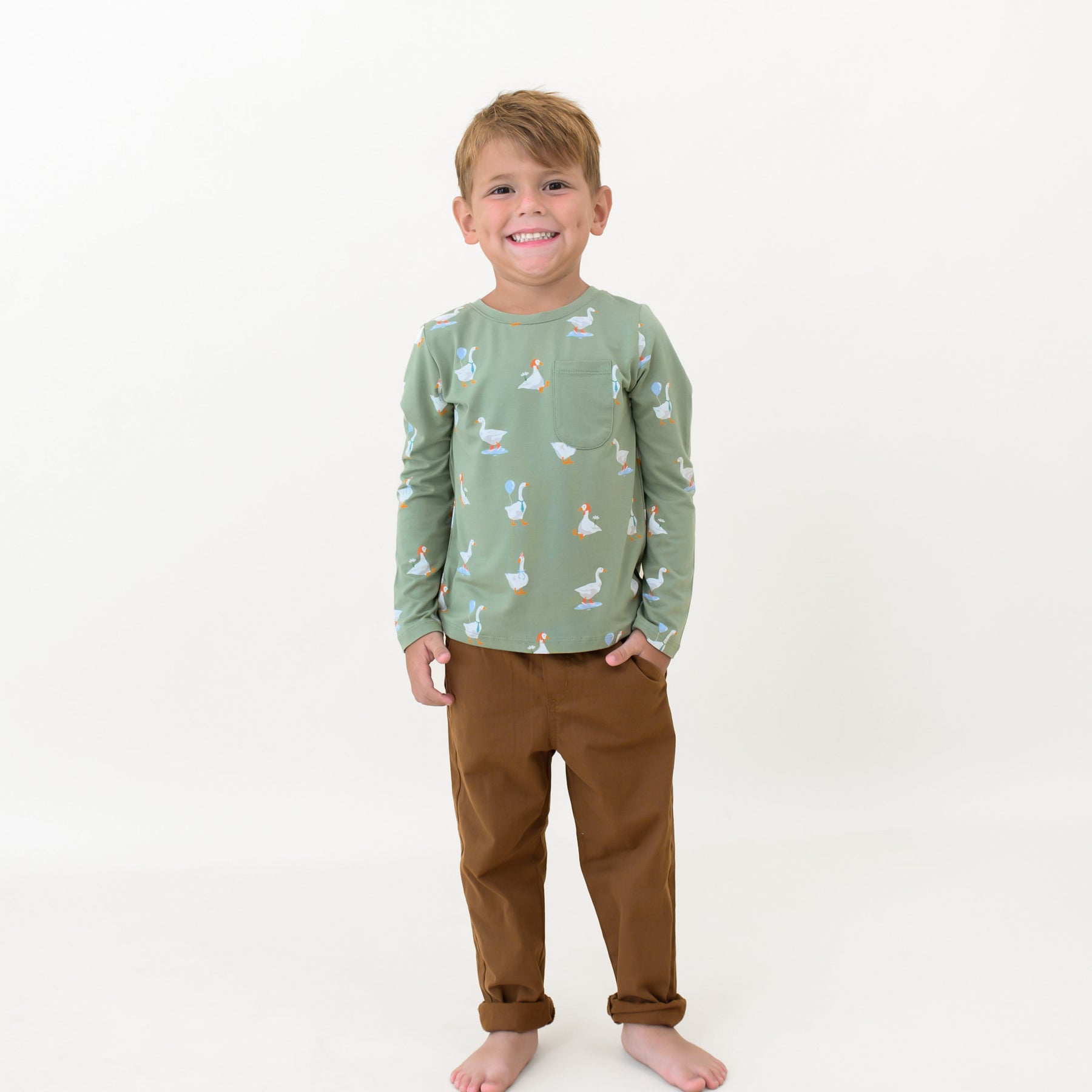 Smiling boy standing with one hand in his pant pocket wearing the Long Sleeve Toddler Crew Neck Tee in Silly Goose in front of  white backdrop