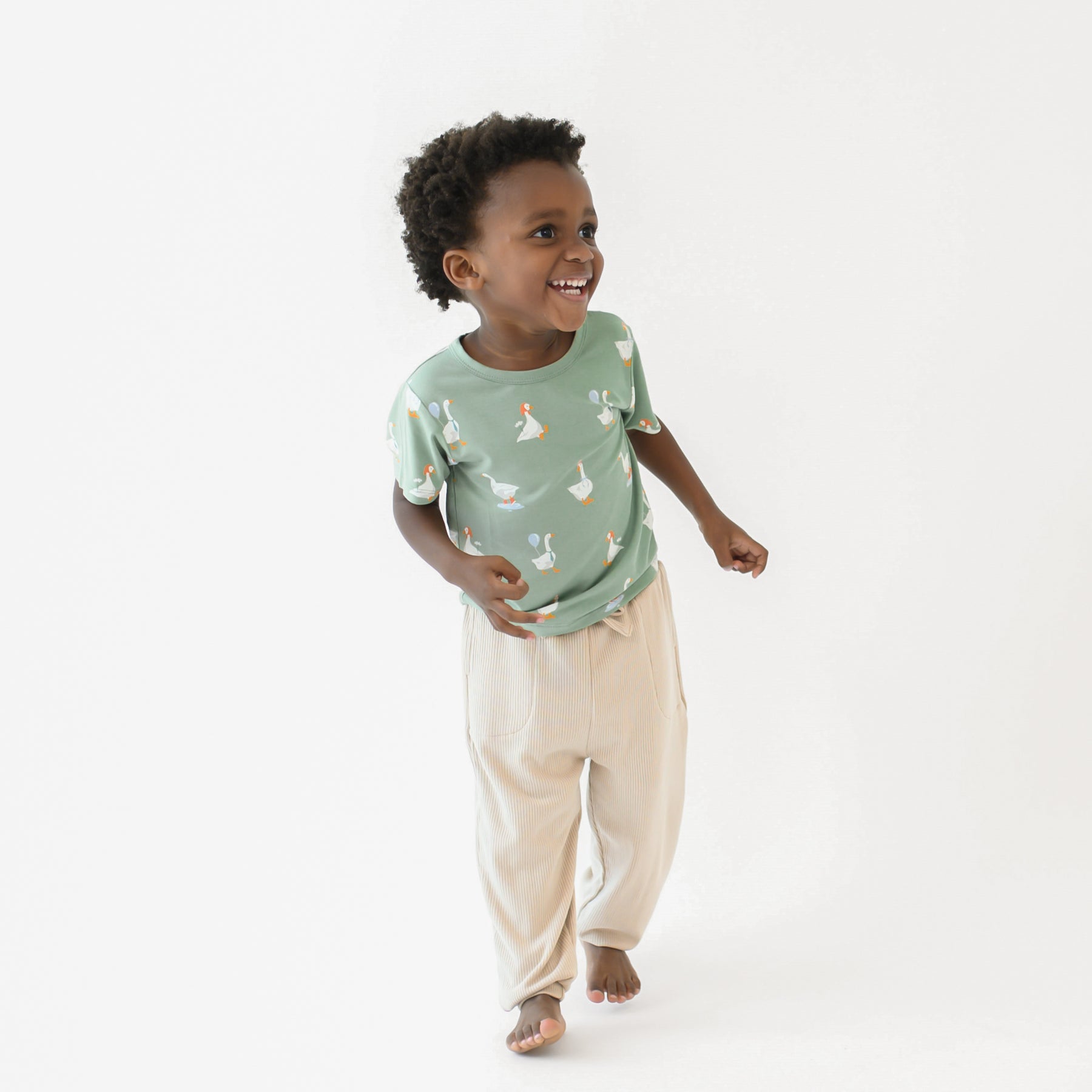 Young boy walking wearing the Toddler Basic Tee in Silly Goose and Bisque Ribbed Jogger pants in front of a white backdrp