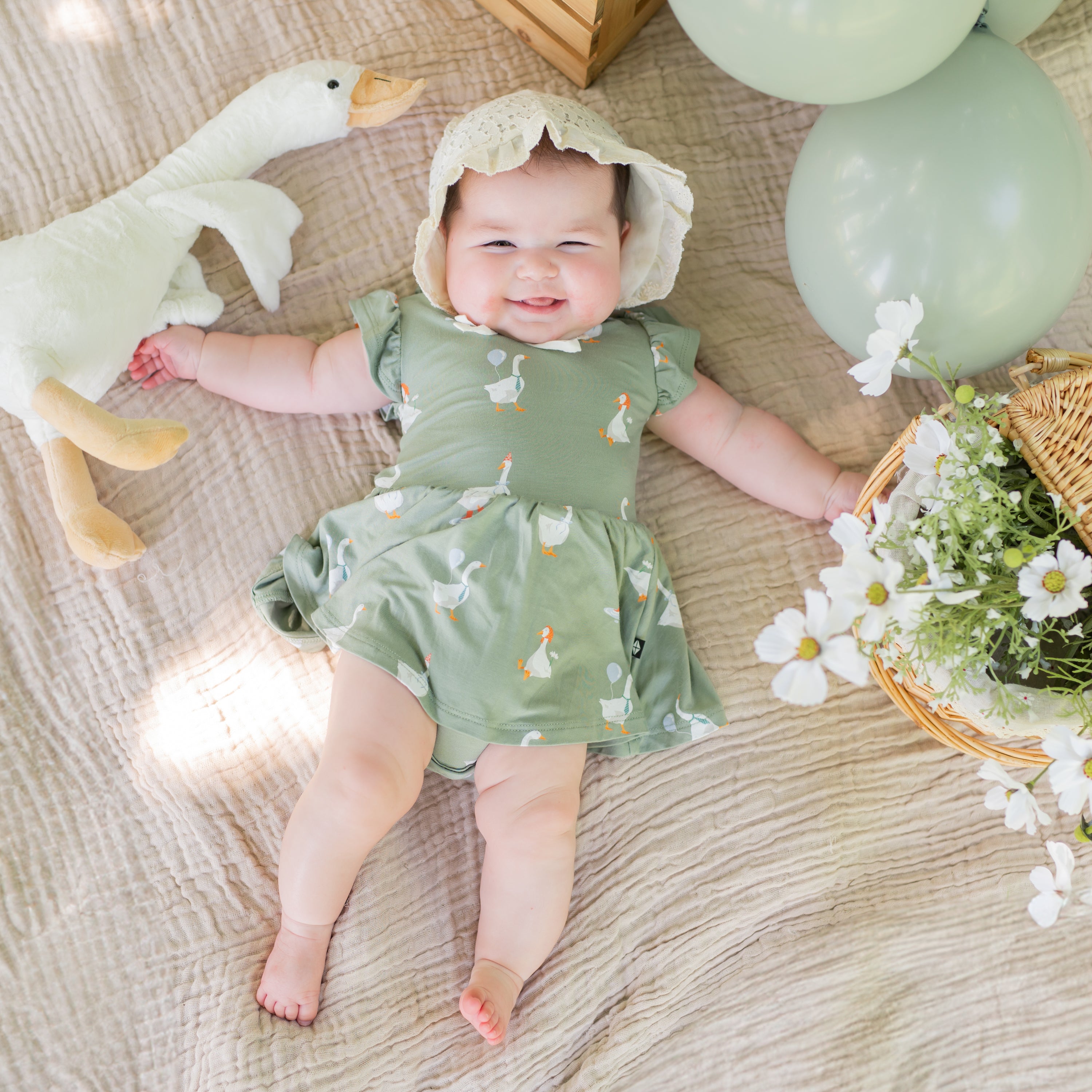 Infant laying in a muslin blanket wearing the Twirl Bodysuit Dress in Silly Goose with linen bonnet surrounded by flowers, balloons and a toy stuffed goose