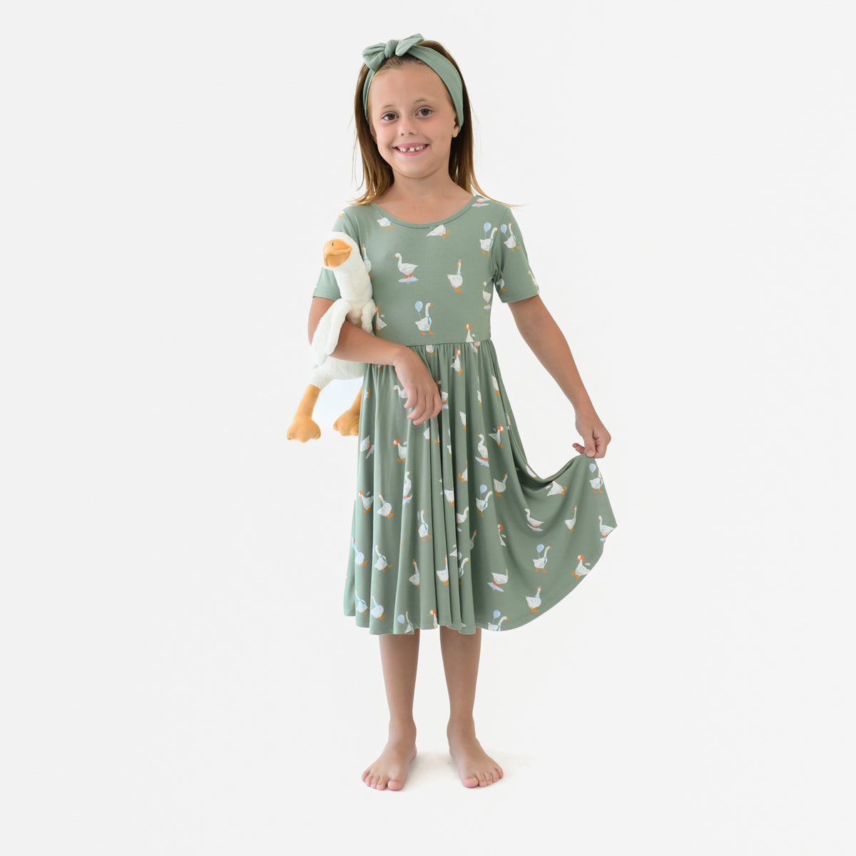 Young girl model standing holding the bottom of the hem of the Twirl Dress in Silly Goose holding a stuffed toy goose