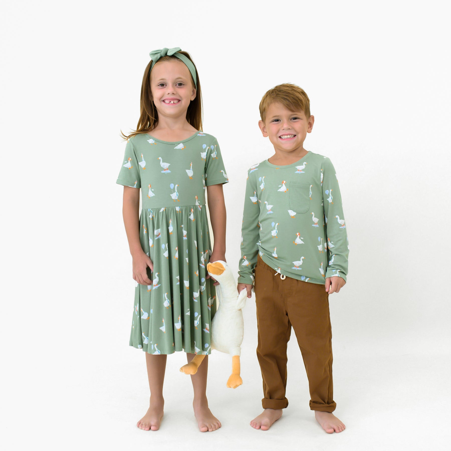 Brother and sister standing side by side holding a toy stuffed goose. Pictured are the Long Sleeve Toddler Crew Neck Tee in Silly Goose and Short Sleeve Twirl dress in Silly Goose