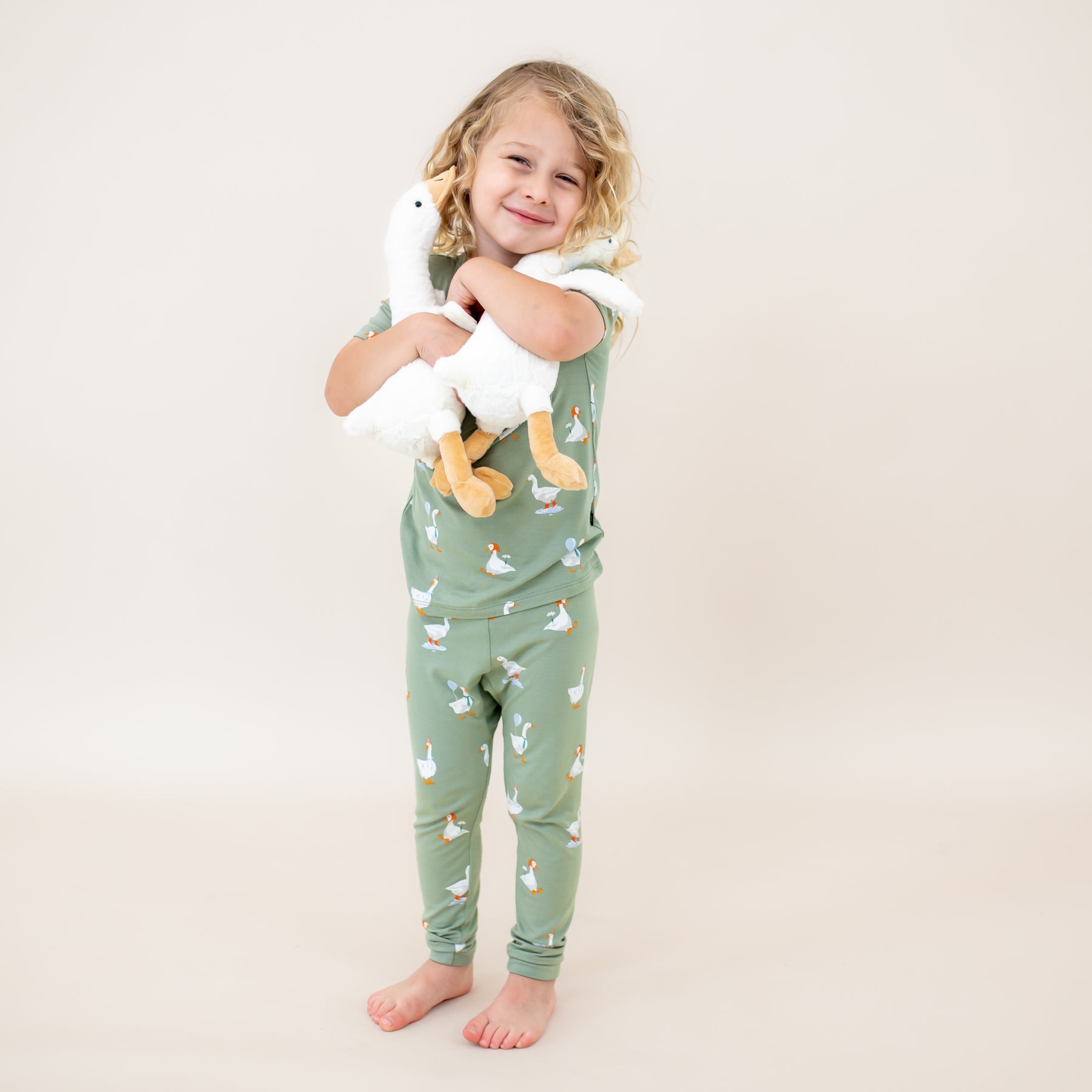 Young child standing holding two toy stuffed geese wearing the Short Sleeve with Pants Pajamas in Silly Goose in front of a light neutral backdrop